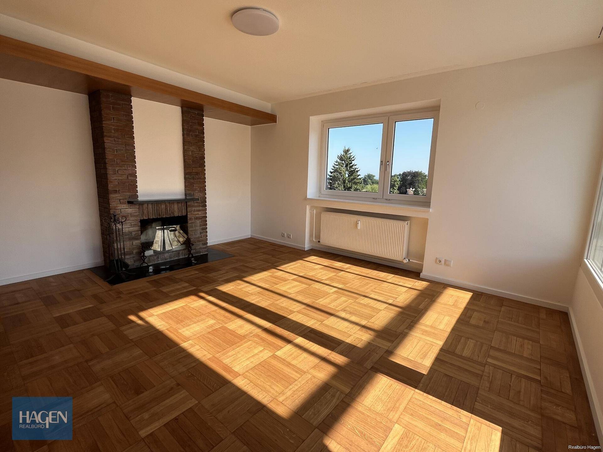 Helles Wohnzimmer mit klassischem Parkettboden, einem gemauerten Kamin und großem Fenster mit Grünblick.