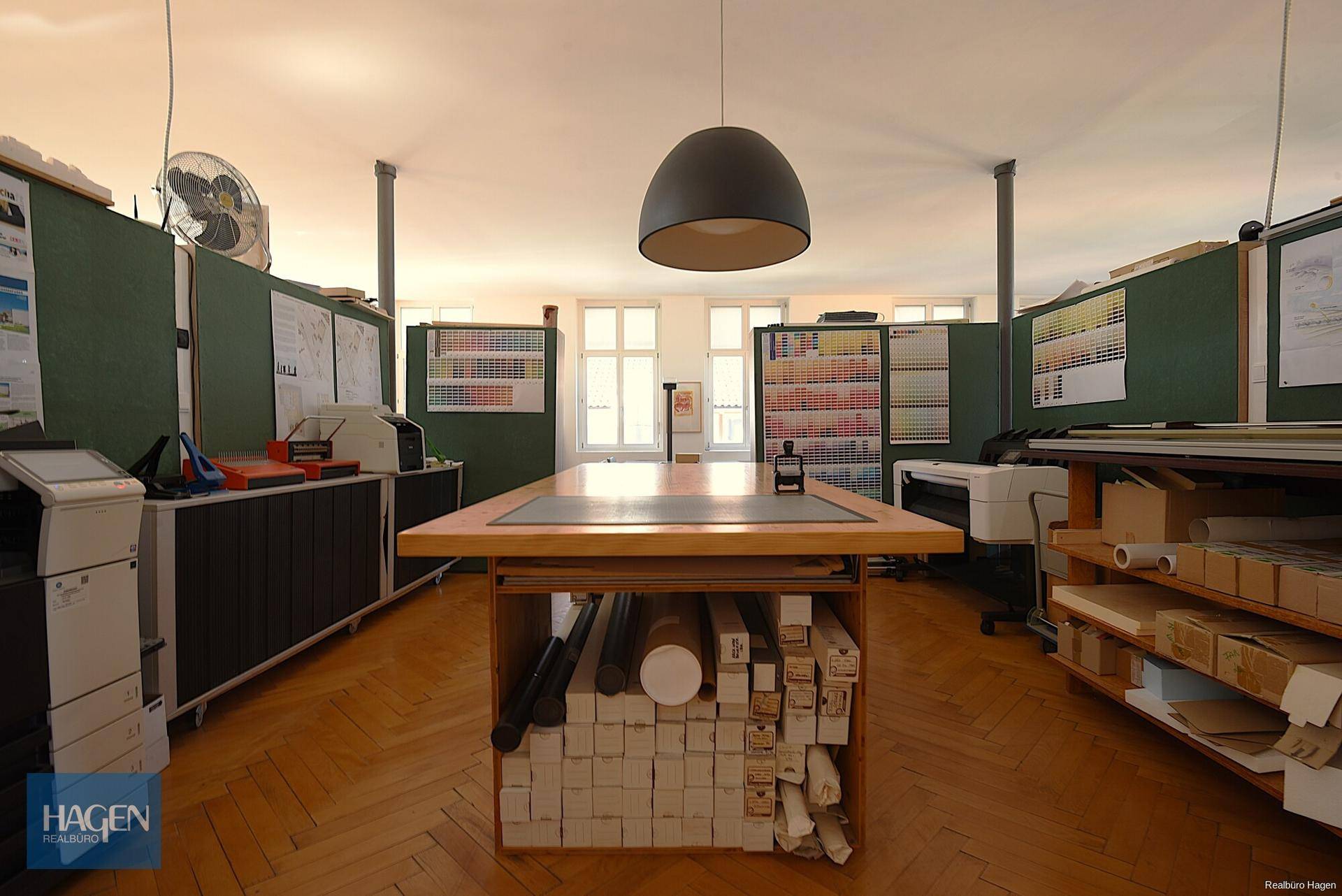 Arbeitsbereich mit Schreibtischen, Regalen und Fischgrätparkett, beleuchtet von einer modernen Hängelampe.