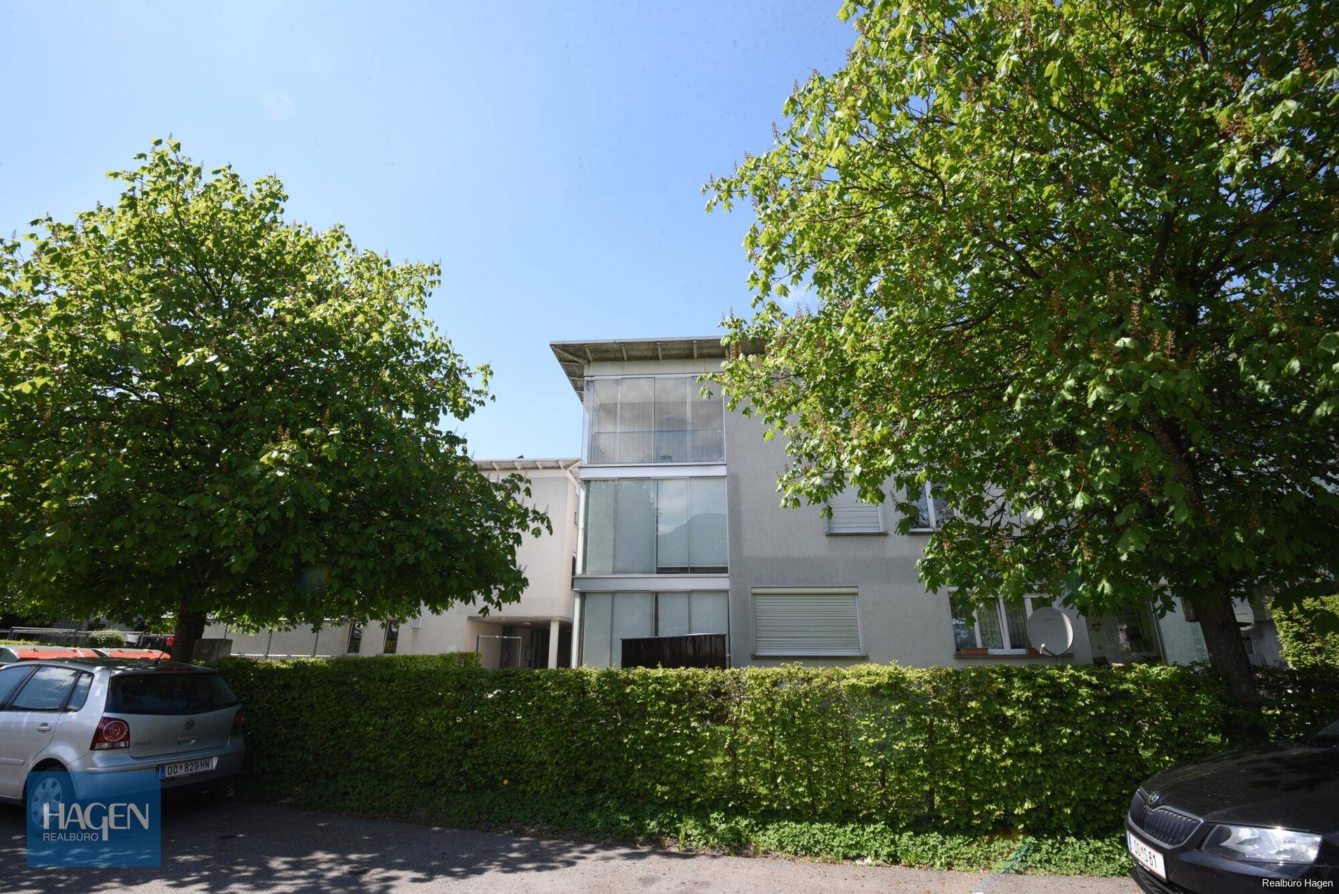 Moderne Gebäudefassade mit großen Fenstern und viel Grün in der Umgebung.