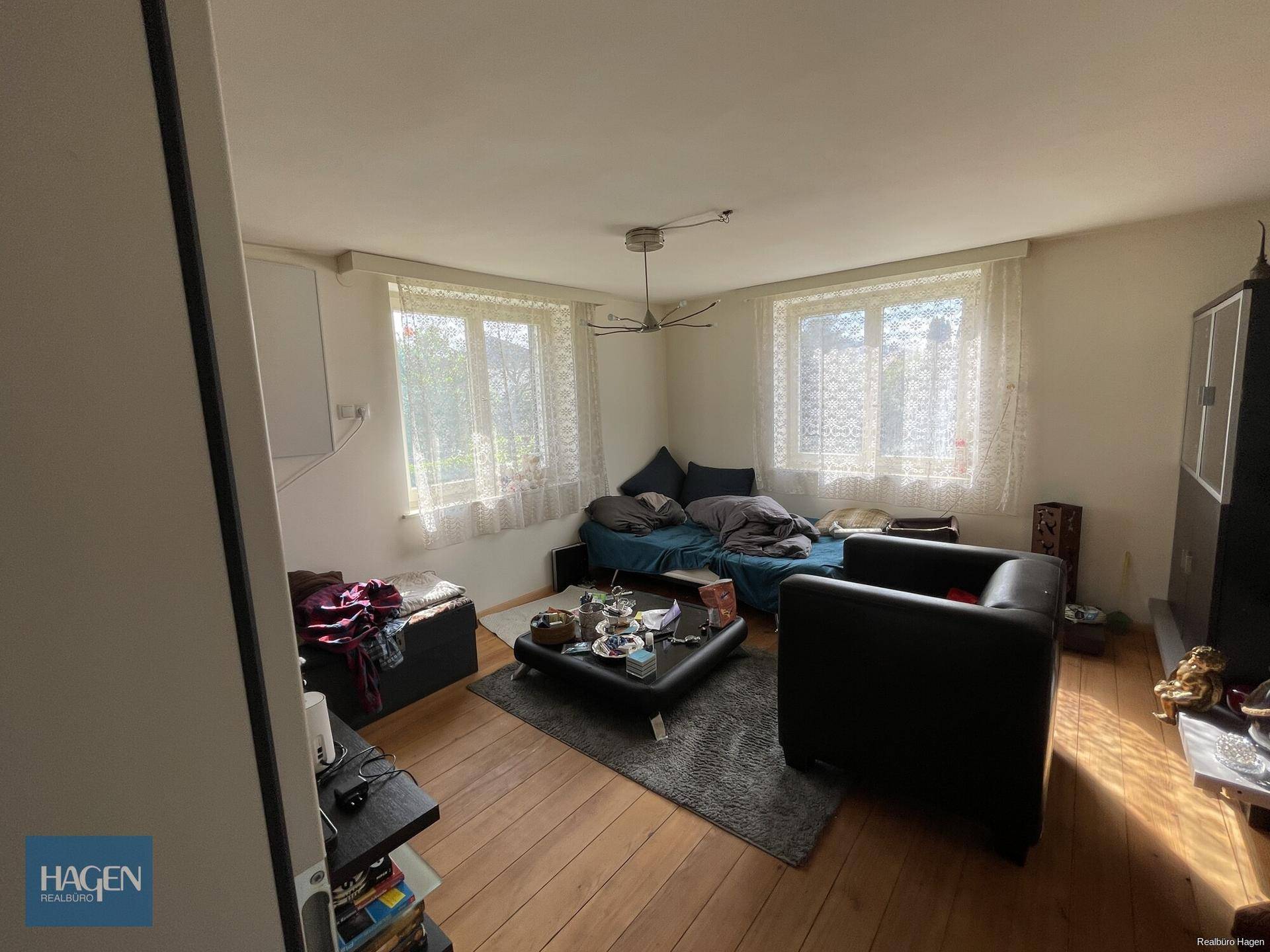 Helles Wohnzimmer mit Holzboden, bequemen Sitzmöbeln und Fenstern mit Spitzenvorhängen.