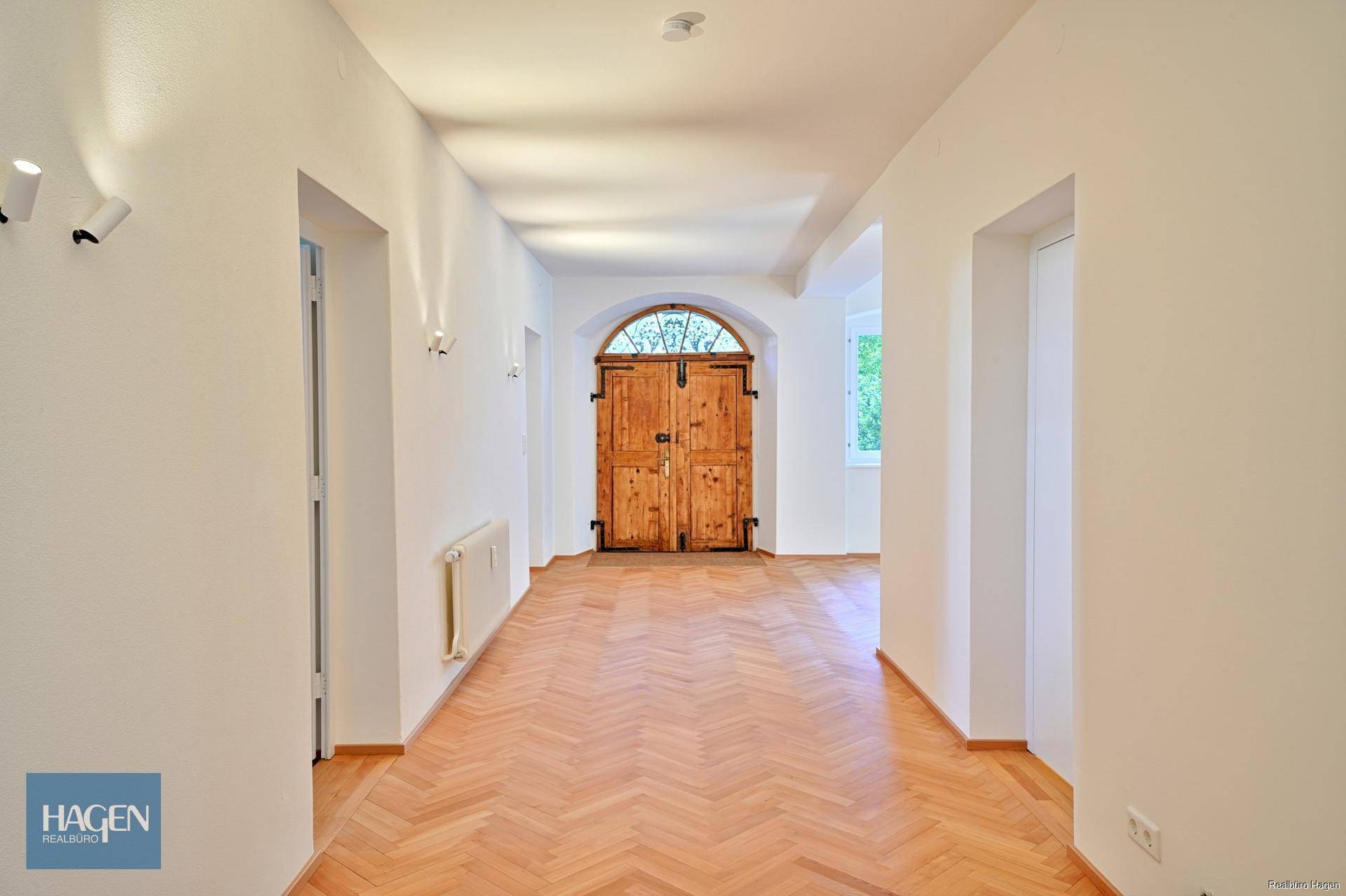Langer Flur mit Fischgrätparkett, weißen Wänden und der großen gewölbten Holztür am Ende.