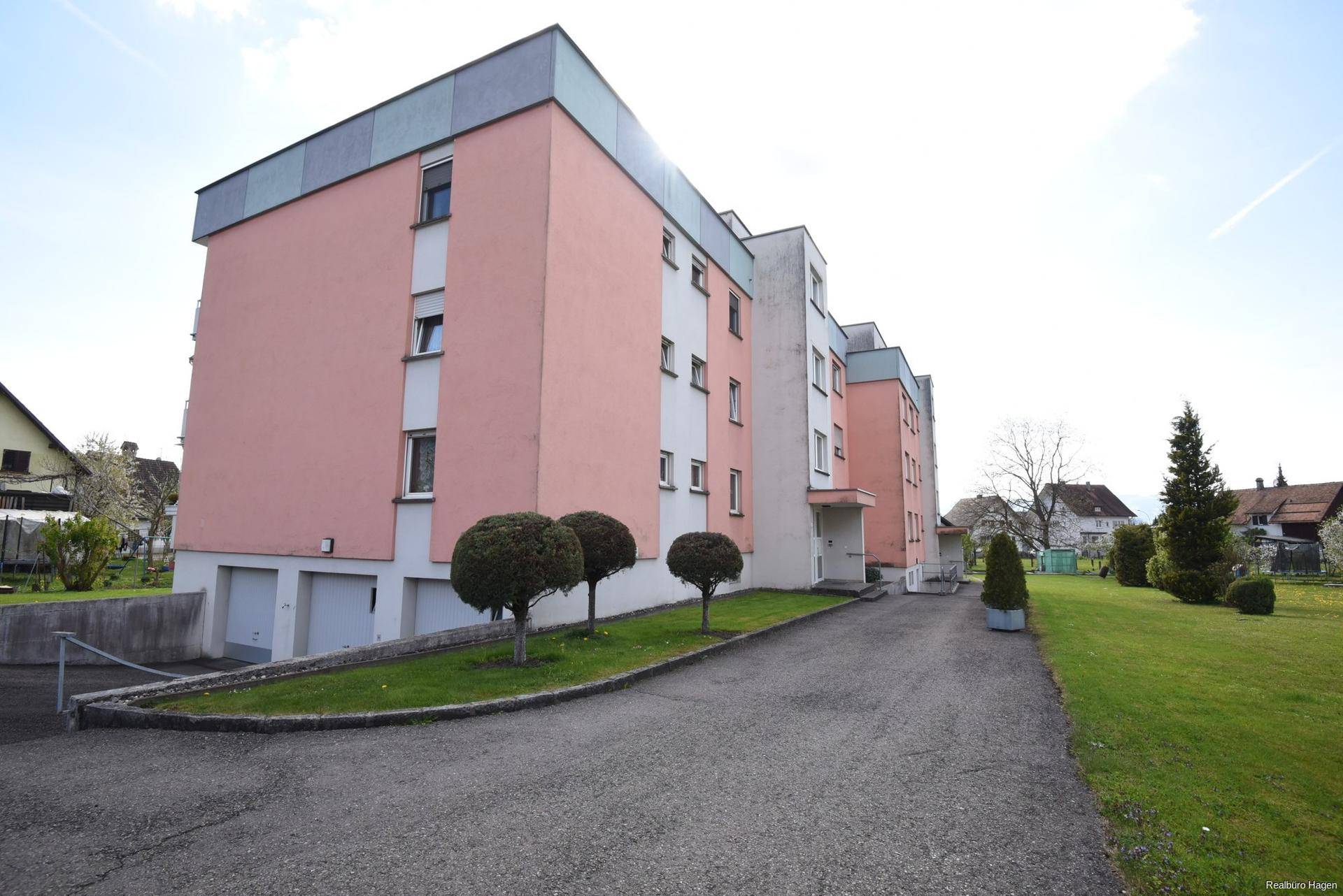Mehrfamilienhaus mit rosa Fassade, Zufahrt und gepflegten Grünflächen in sonniger Lage.