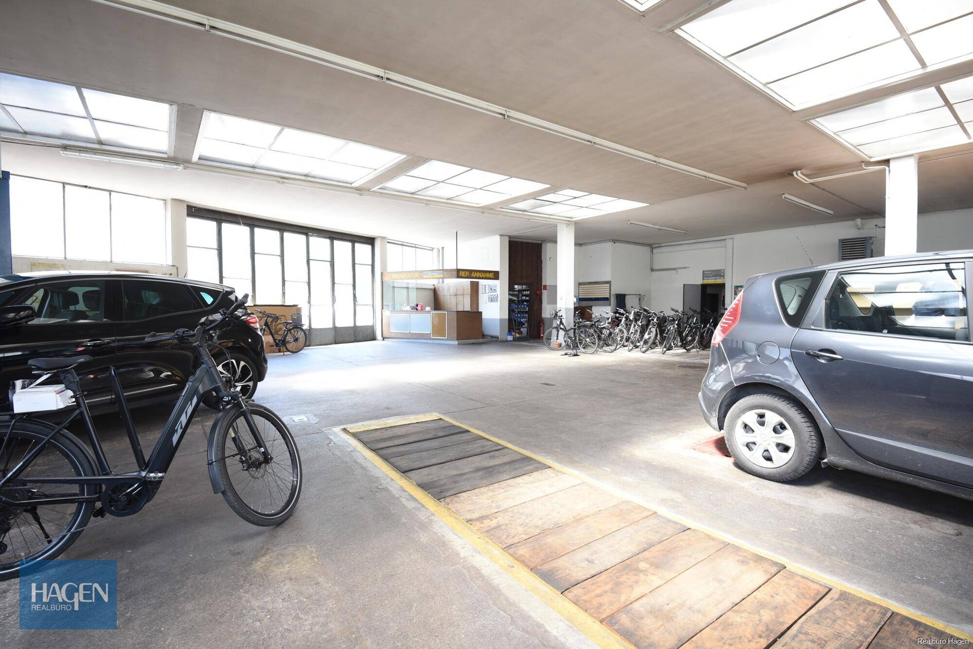 Werkstatt- oder Garagenbereich mit Autos, Fahrrädern, großen Fenstern und Oberlichtern, Betonboden.
