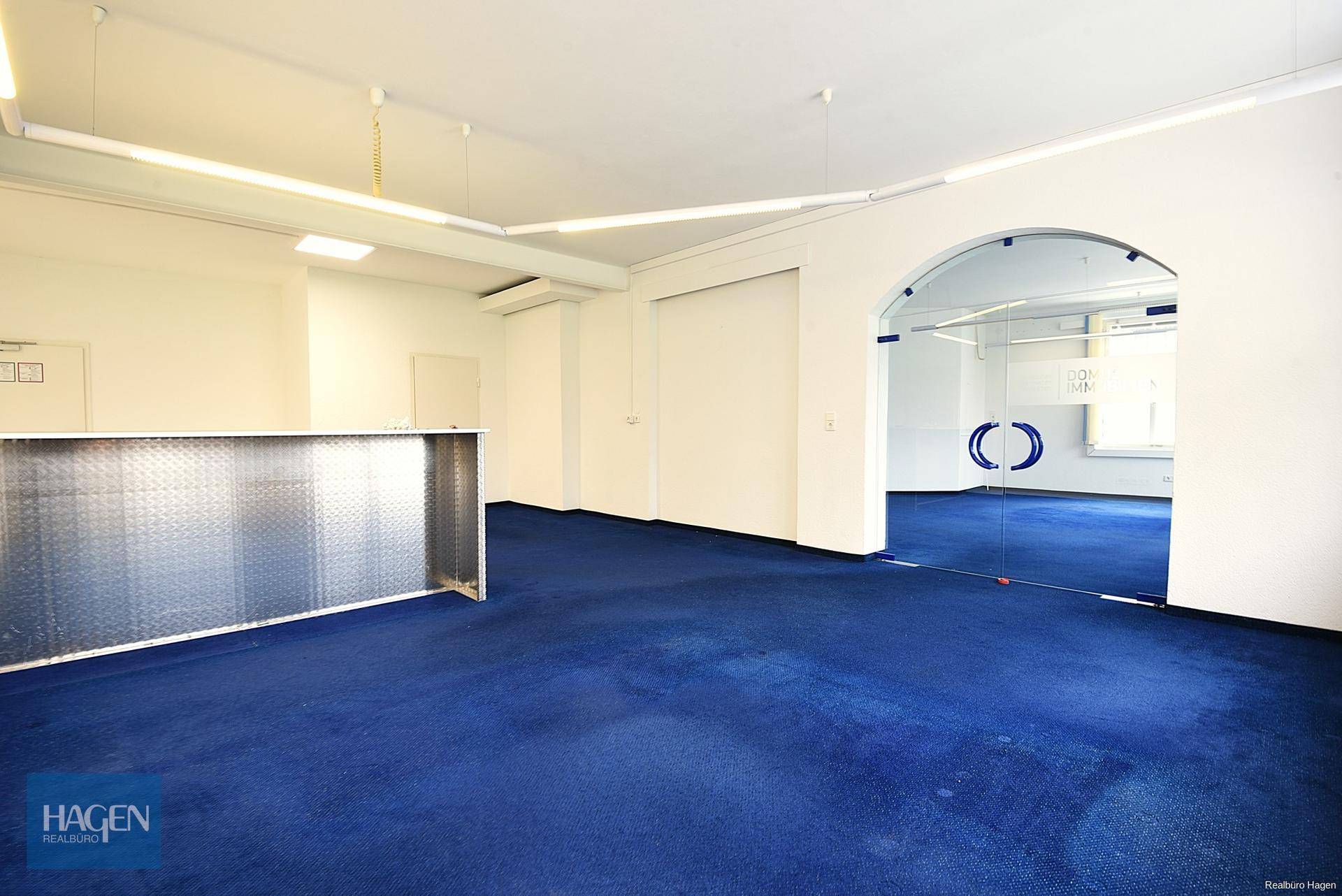 Großer offener Büroraum mit blauem Teppichboden und einer modernen Glastür zu einem weiteren Bereich.
