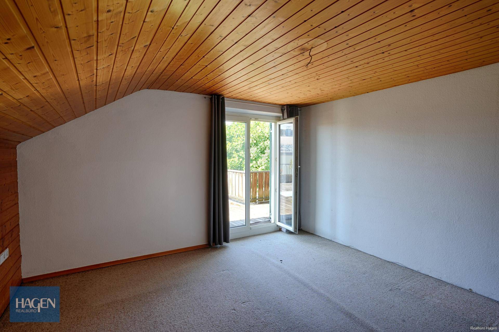 Dachgeschosszimmer mit Holzdecke, Teppichboden und Zugang zum Balkon.