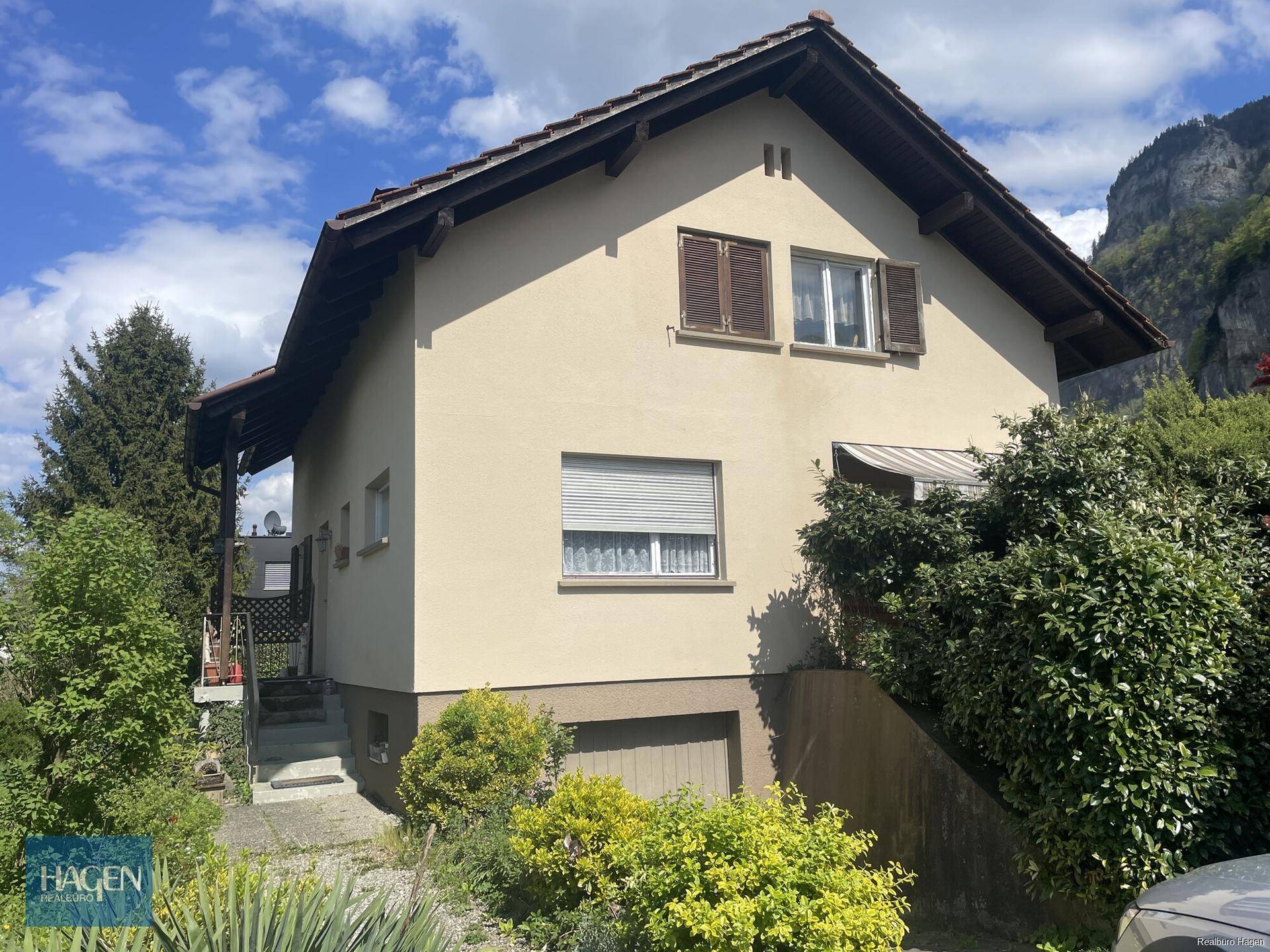 Seitenansicht des Hauses mit Garagenzufahrt und Treppe, im Hintergrund ist ein Berg sichtbar.