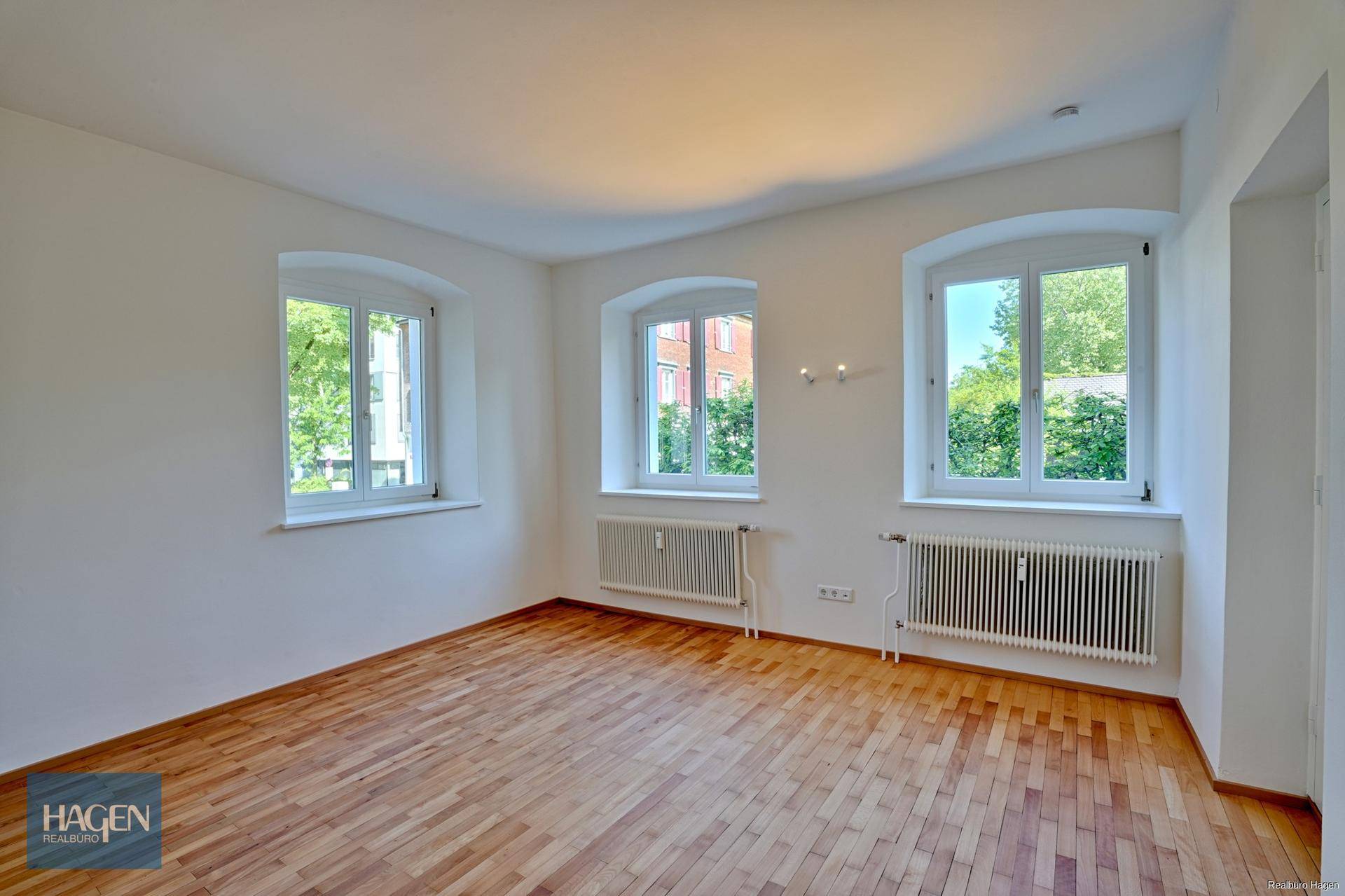 Leeres Zimmer mit Fischgrätparkett, weißen Wänden und drei Fenstern mit Blick ins Grüne.