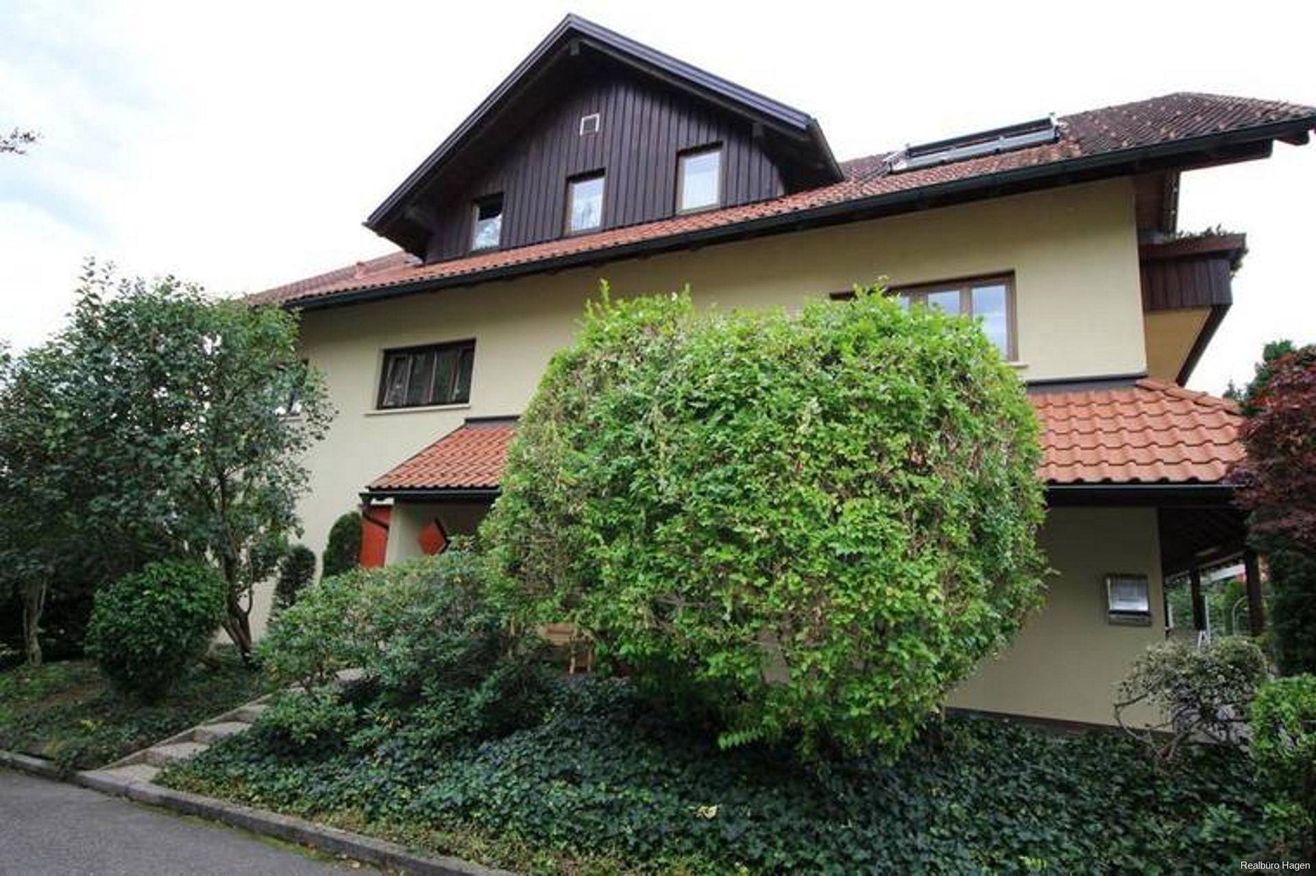 Traditionelles Wohnhaus mit beiger Fassade, braunem Dach und gepflegter Gartenanlage, umgeben von üppigem Grün.