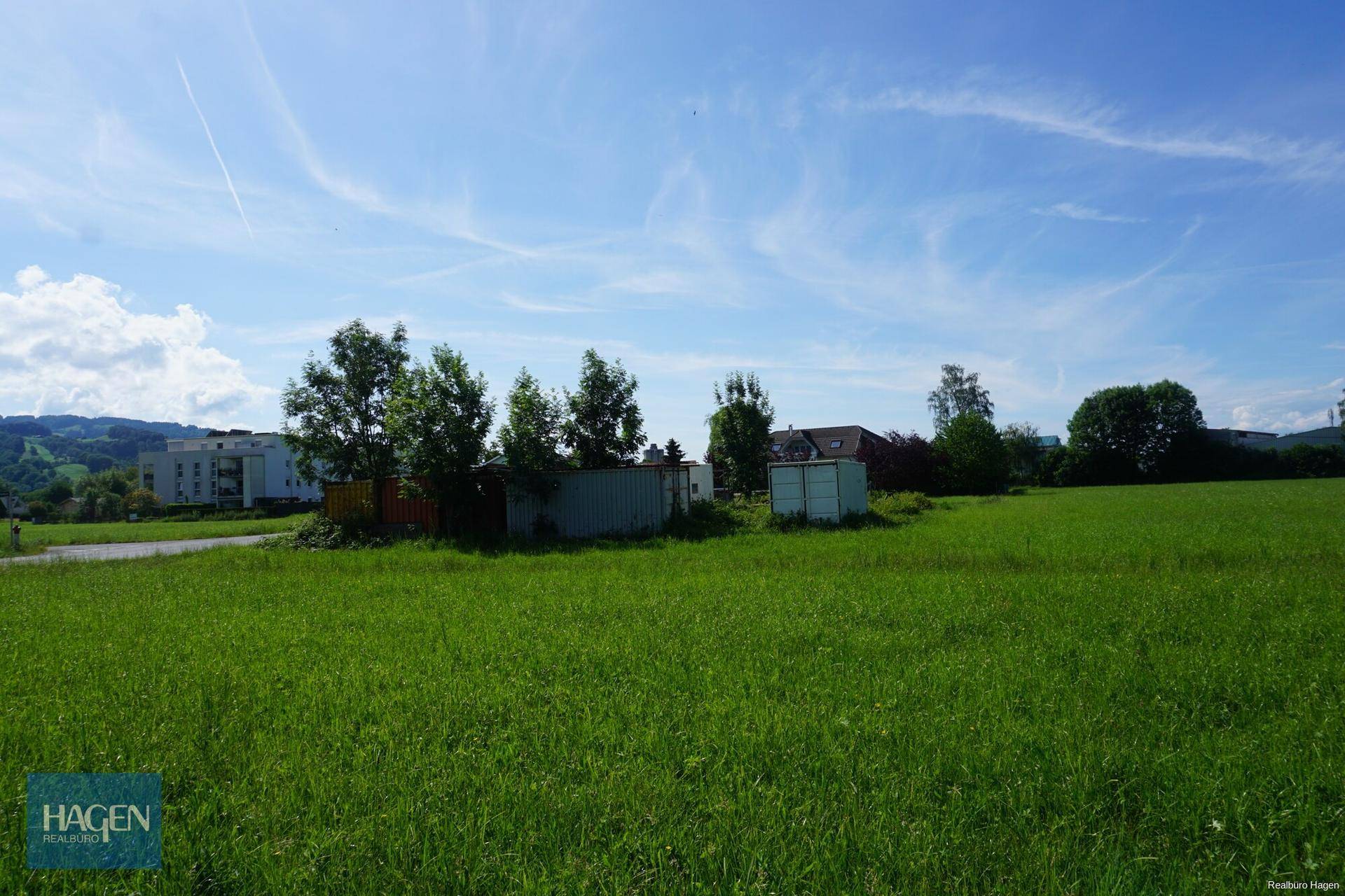 Offene grüne Fläche mit kleinen Schuppen und Wohngebäuden in der Ferne unter einem weiten Himmel.