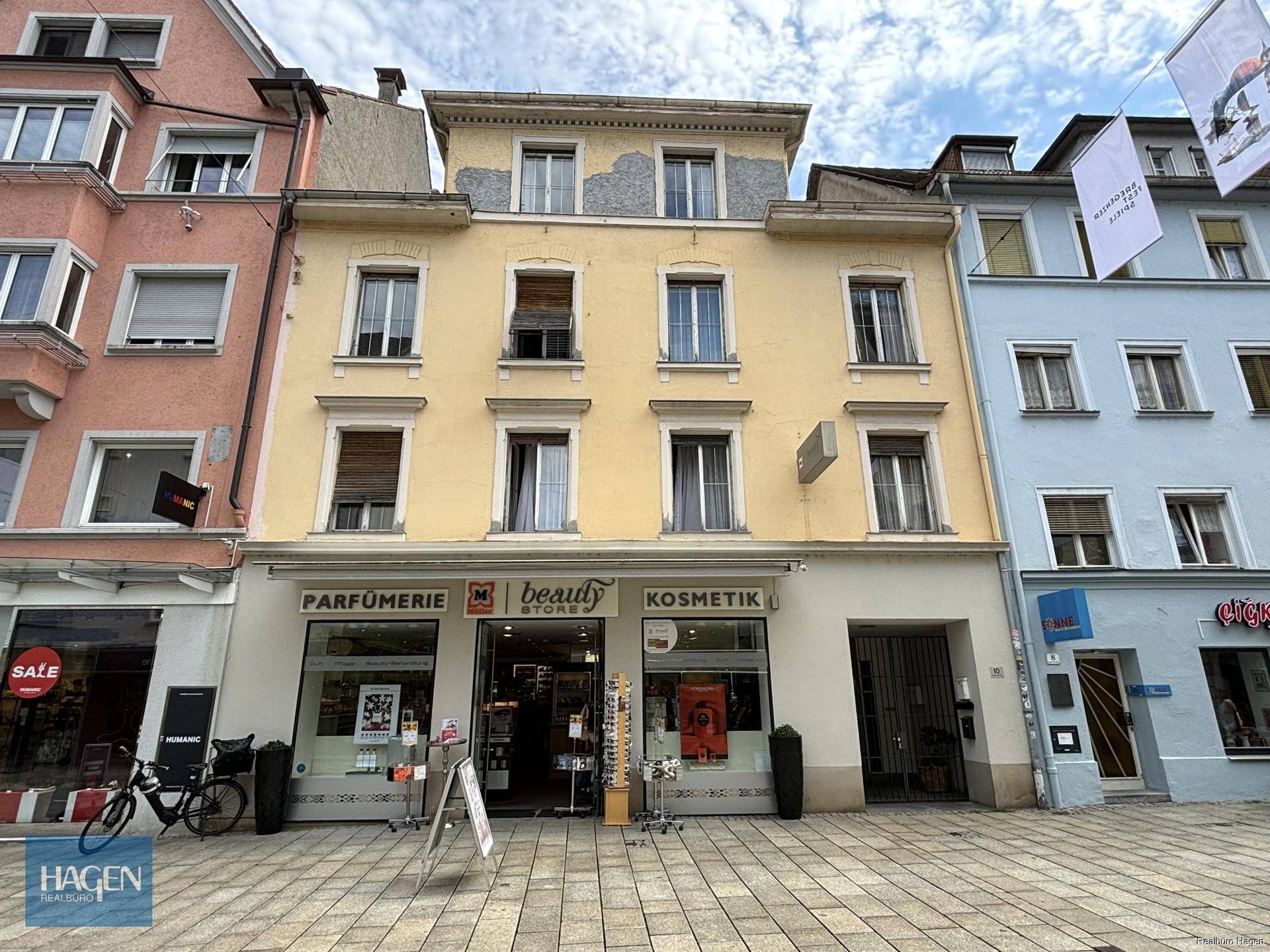 Gelbe Fassade eines Mehrfamilienhauses mit Geschäften im Erdgeschoss an einer belebten Straße.