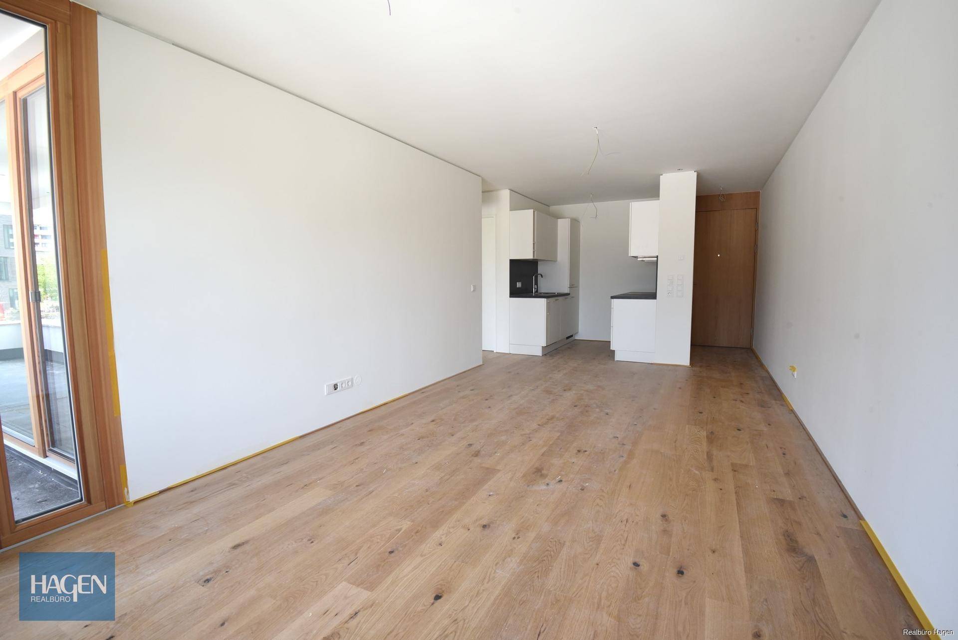 Heller, leerer Wohnbereich mit Holzboden und Zugang zu einem Balkon oder einer Terrasse.