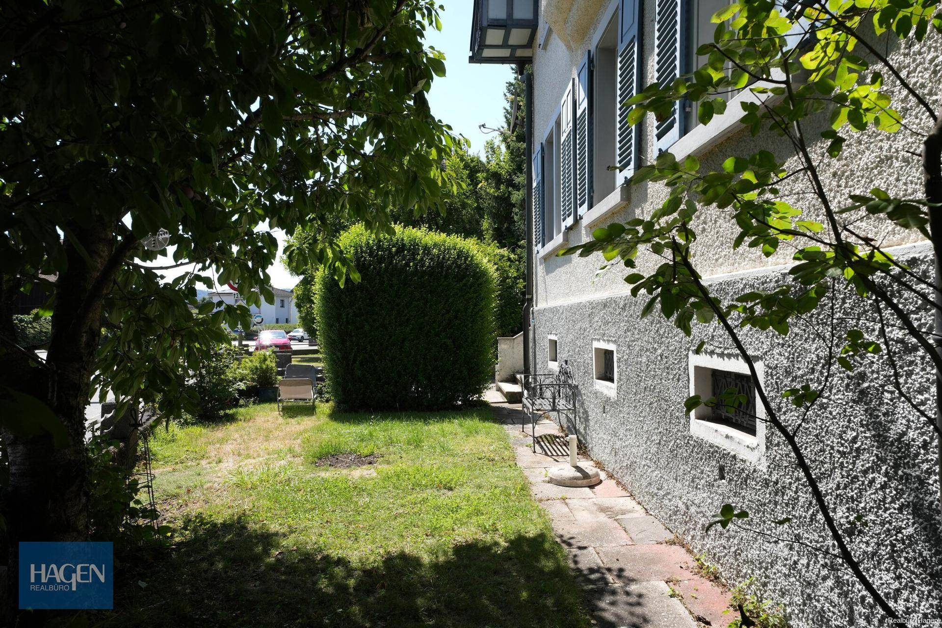 Seitlicher Gartenbereich mit gepflegtem Rasen, Hecke und schattenspendenden Bäumen.