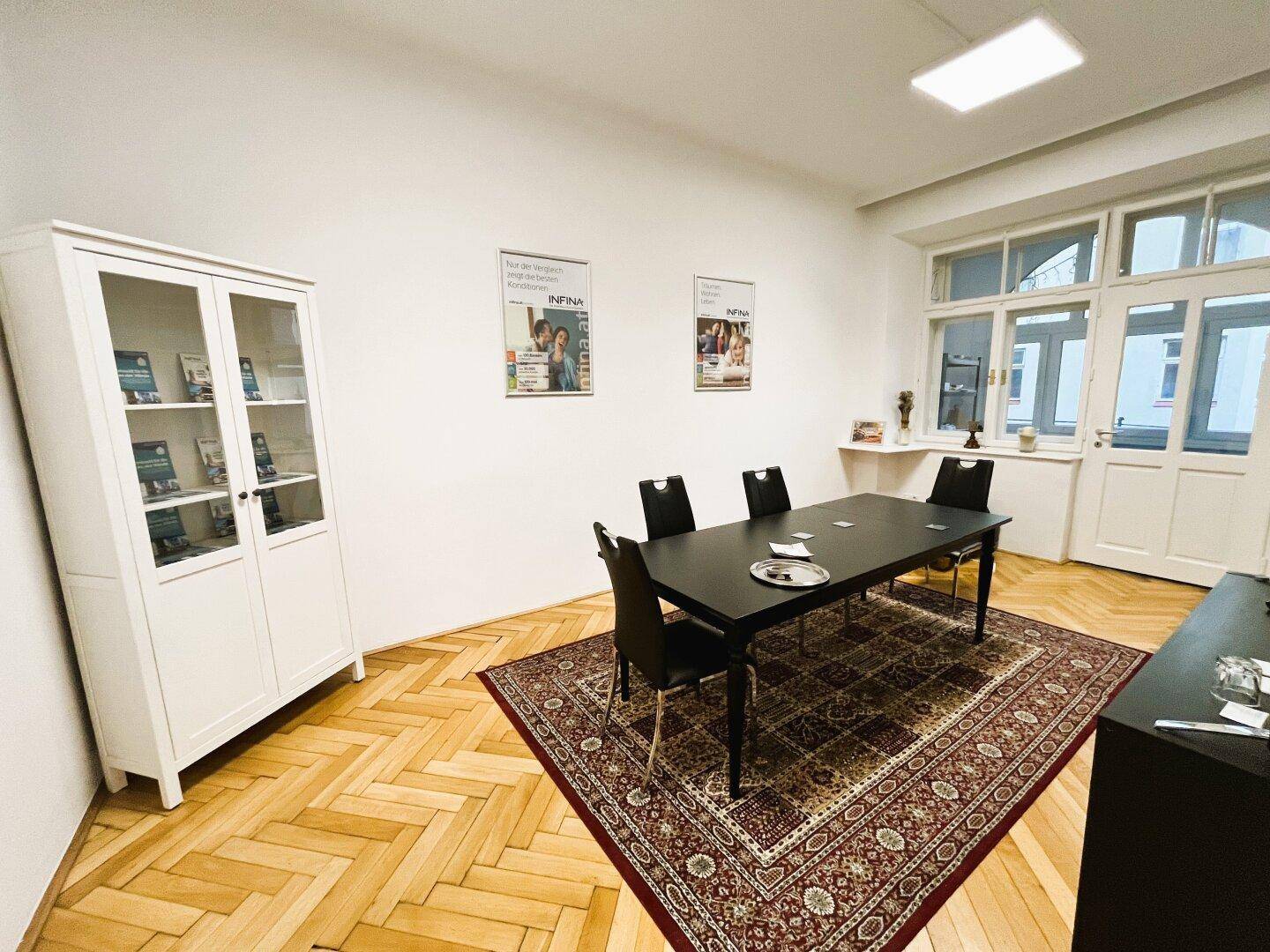 Heller Büroraum mit Fischgrätparkett, einem großen Tisch und einem Vitrinenschrank am Fenster.
