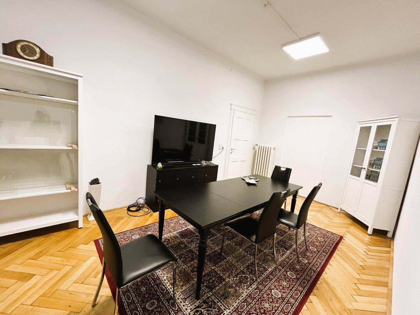 Büroraum mit Fischgrätparkett, einem großen Tisch, Stühlen und einem Fernseher an der Wand.