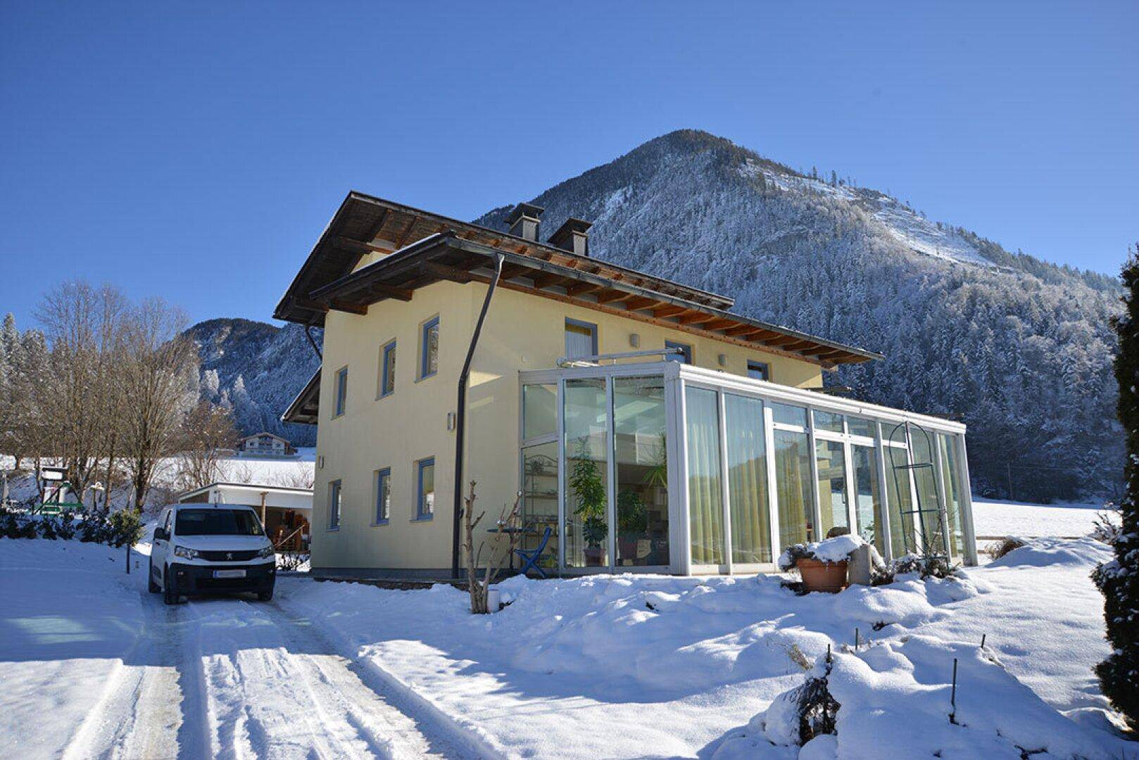 Hausfassade mit großem verglastem Wintergarten und verschneiter Berglandschaft im Hintergrund.