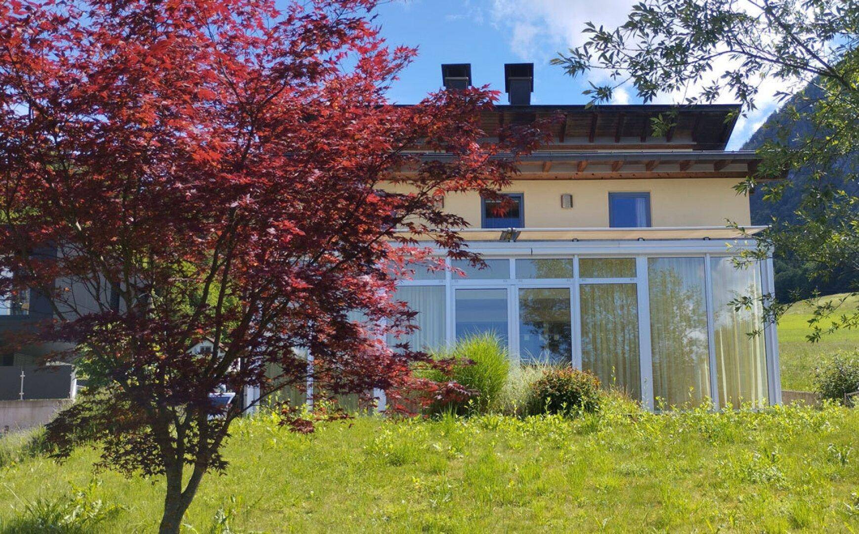 Hausfassade mit Wintergarten, umgeben von Bäumen und einem auffälligen roten Baum im Vordergrund.