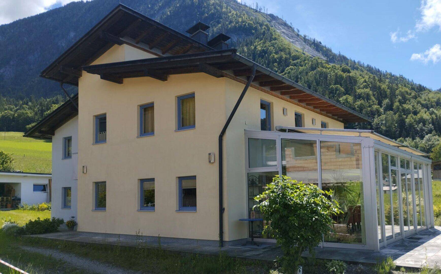 Seitenansicht des Hauses mit Wintergarten und grüner Berglandschaft im Hintergrund.