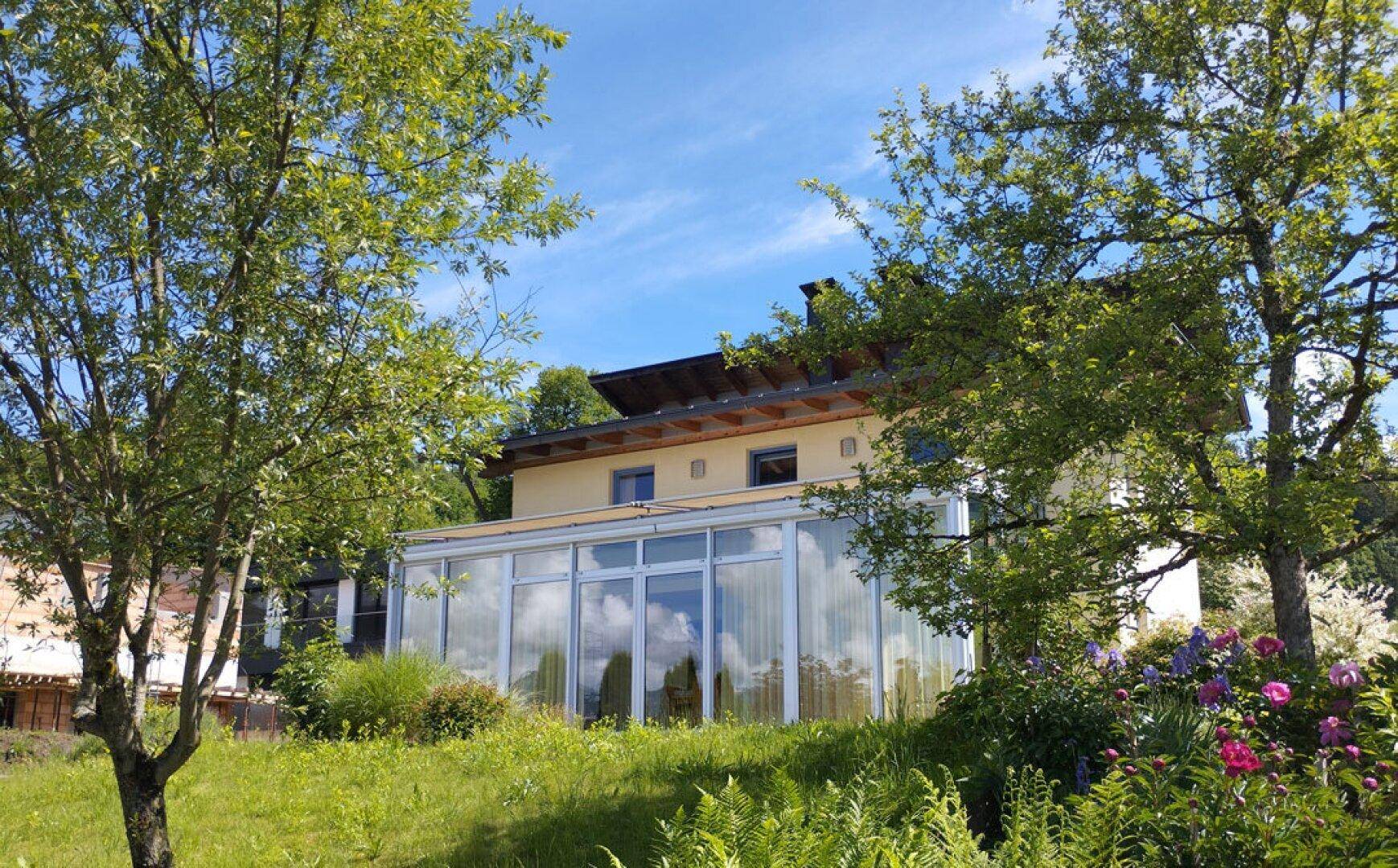 Hausfassade mit großem Wintergarten, umgeben von üppigem Grün und Bäumen unter blauem Himmel.