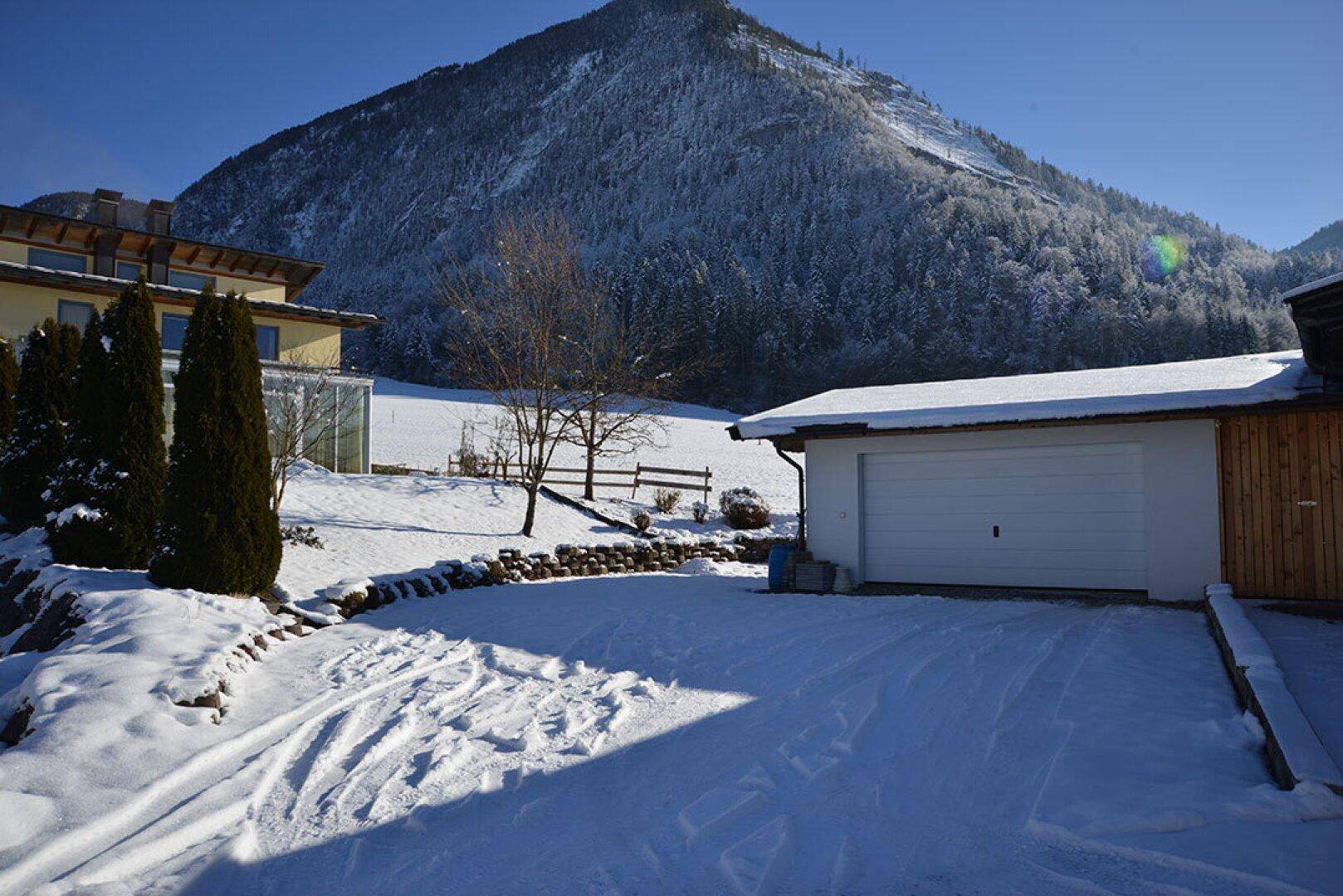 Garage mit schneebedeckter Einfahrt und verschneiter Berglandschaft im Hintergrund, ideal für Fahrzeuge.