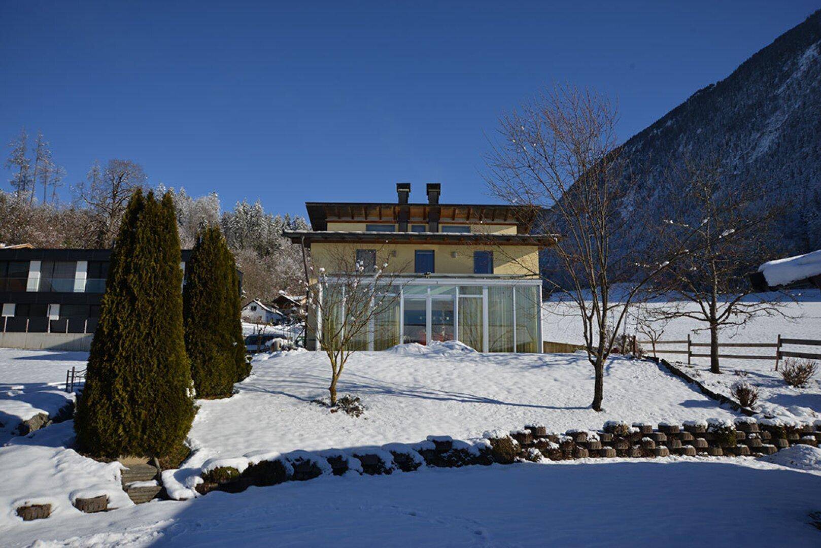 Hausfassade im Winter, umgeben von einem schneebedeckten Garten und Bäumen.