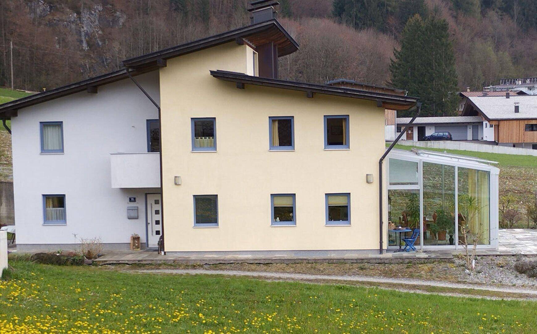 Vorderansicht des Hauses mit Wintergarten in ländlicher Umgebung und Bergen im Hintergrund.