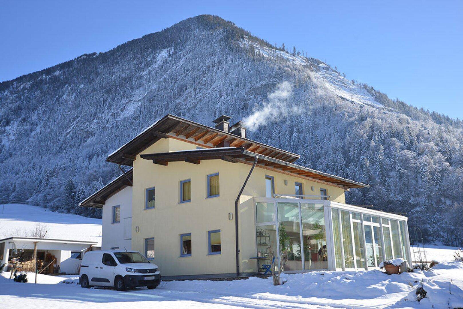 Hausfassade im Winter mit Schnee, umgeben von verschneiten Bergen und einem Wintergarten.