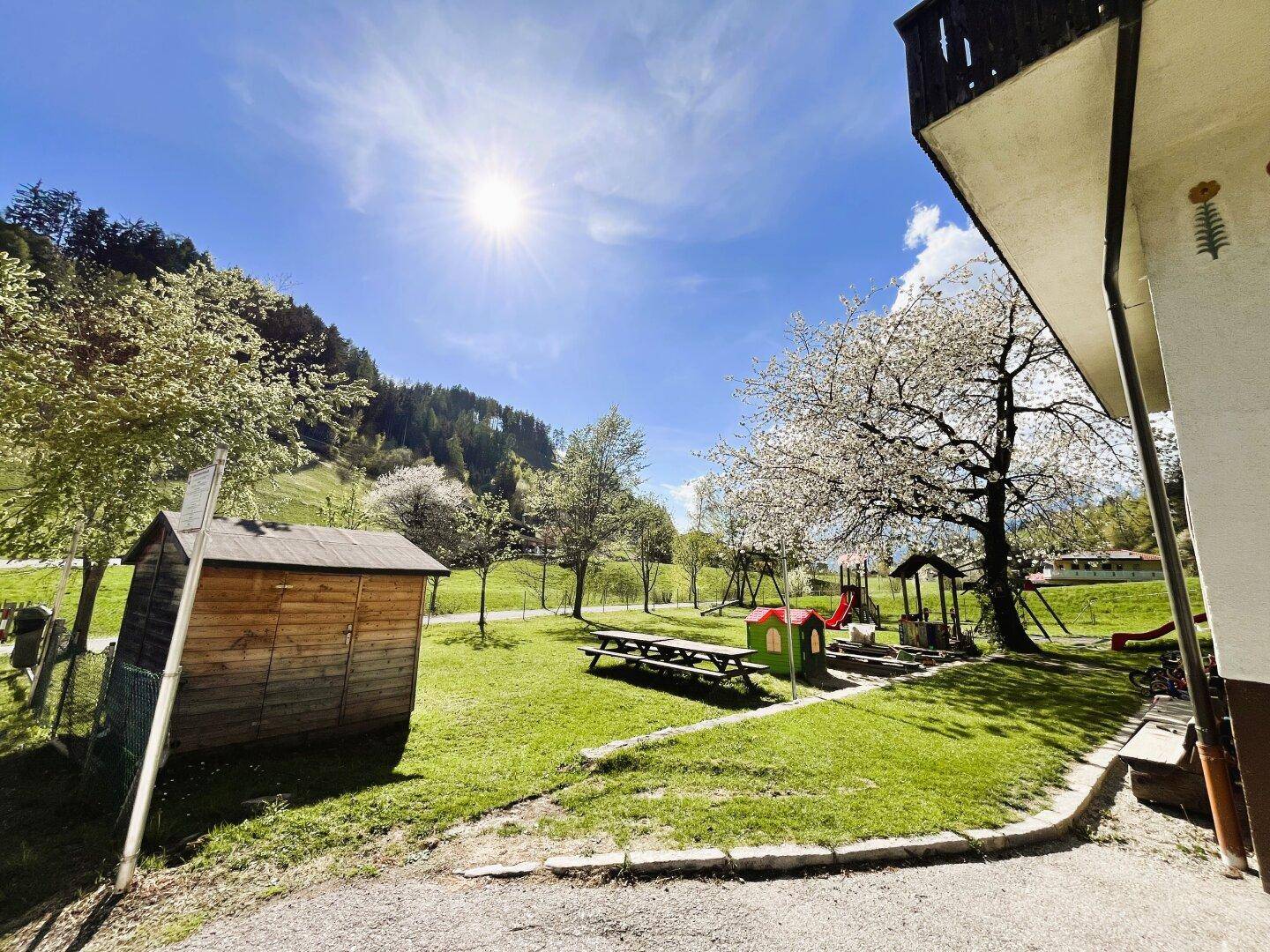 Grüner Gartenbereich mit Kinderspielplatz, Schaukeln und einer kleinen Gartenhütte unter blauem Himmel.