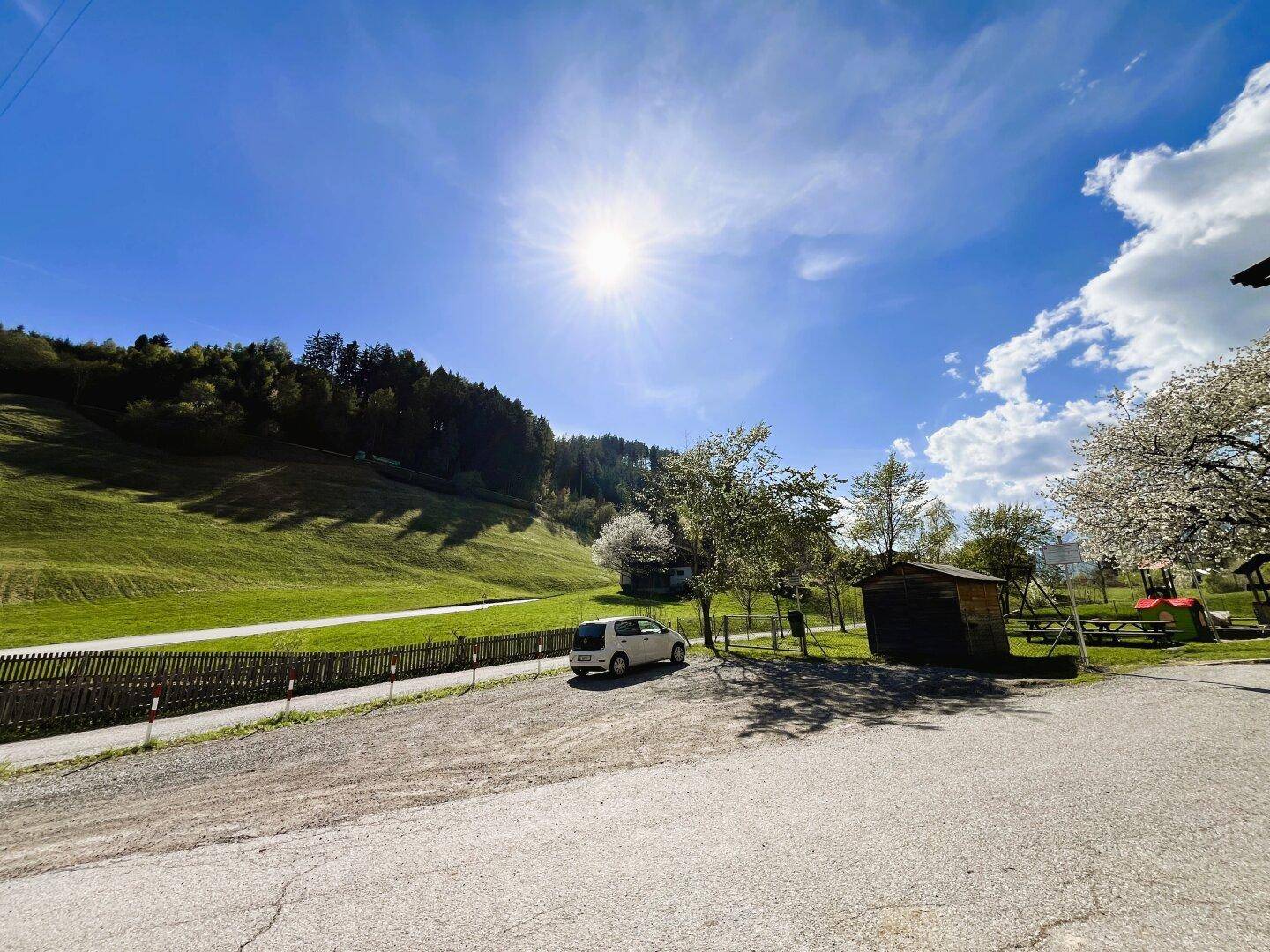 Sonniger Außenbereich mit Parkplatz, Holzschuppen und Blick auf grüne Wiesen und Berge.