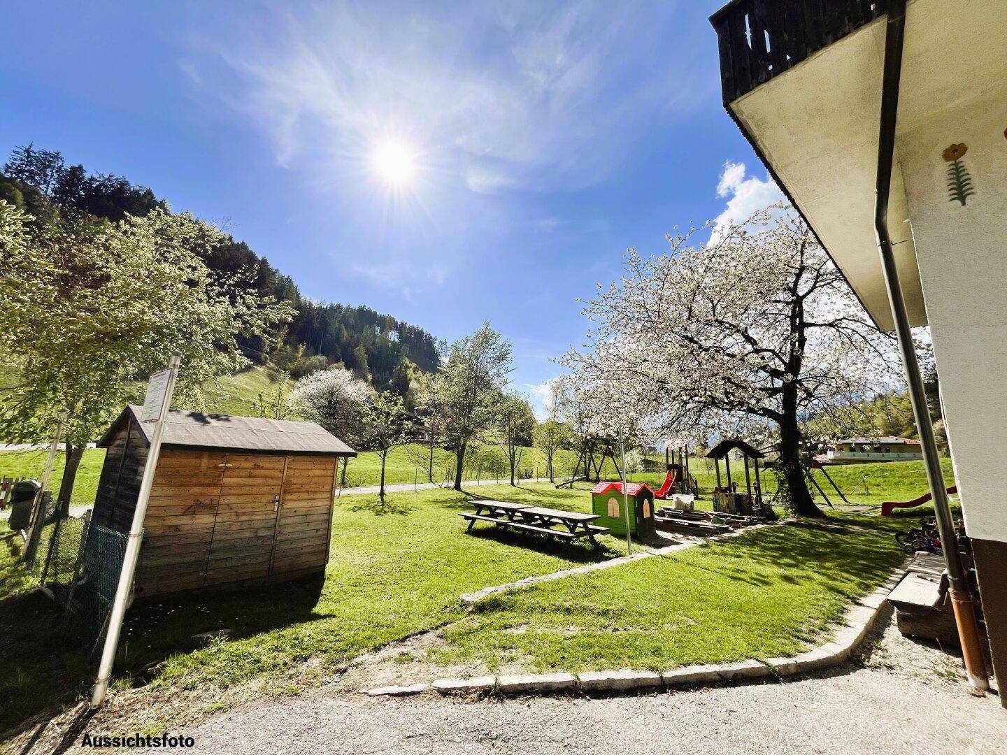 Heller Außenbereich mit Spielplatz, Holzschuppen und blühenden Bäumen unter blauem Himmel.