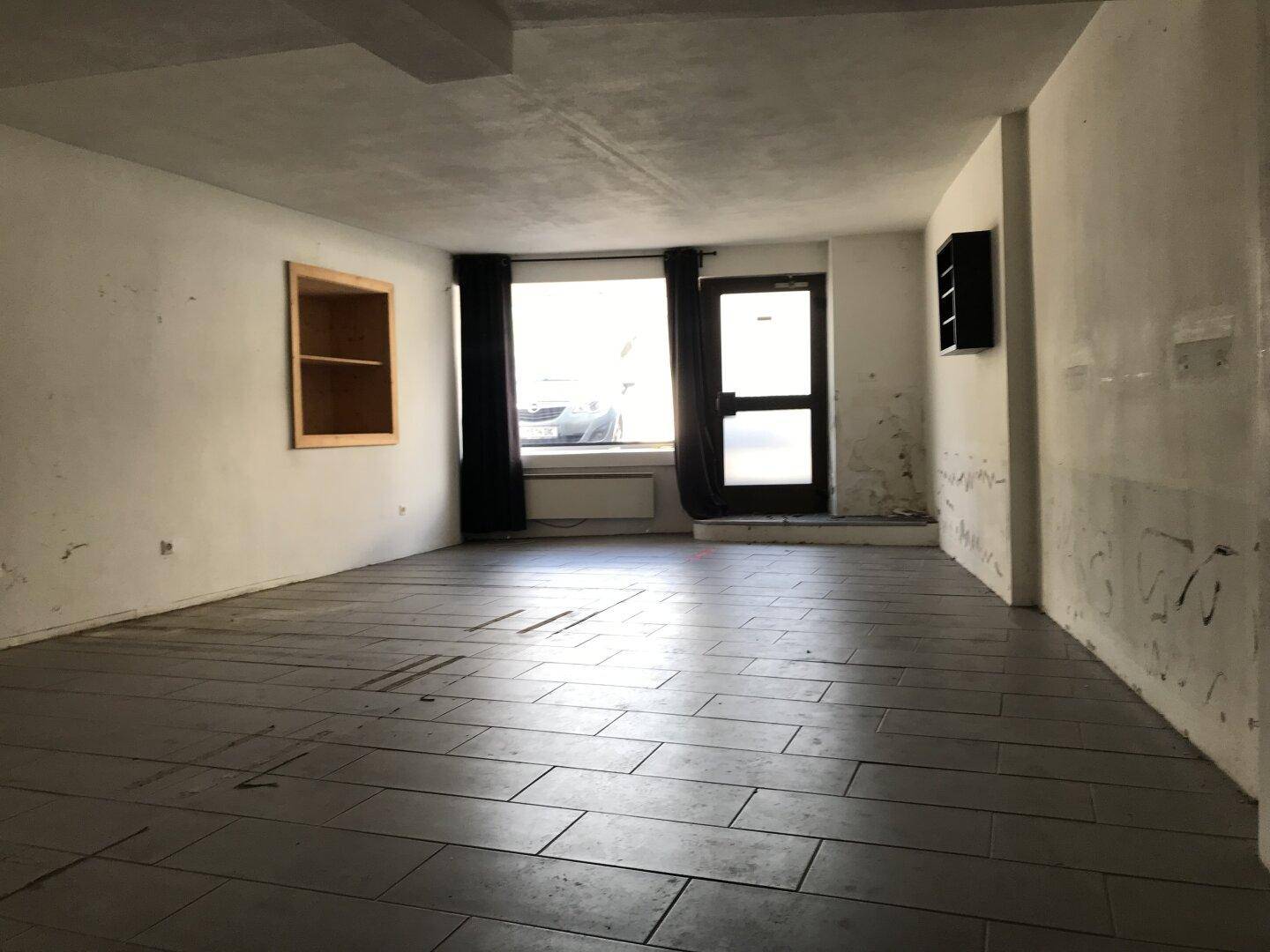 Geräumiger, leerer Raum mit dunklen Fliesen und einem Fenster zur Straße.