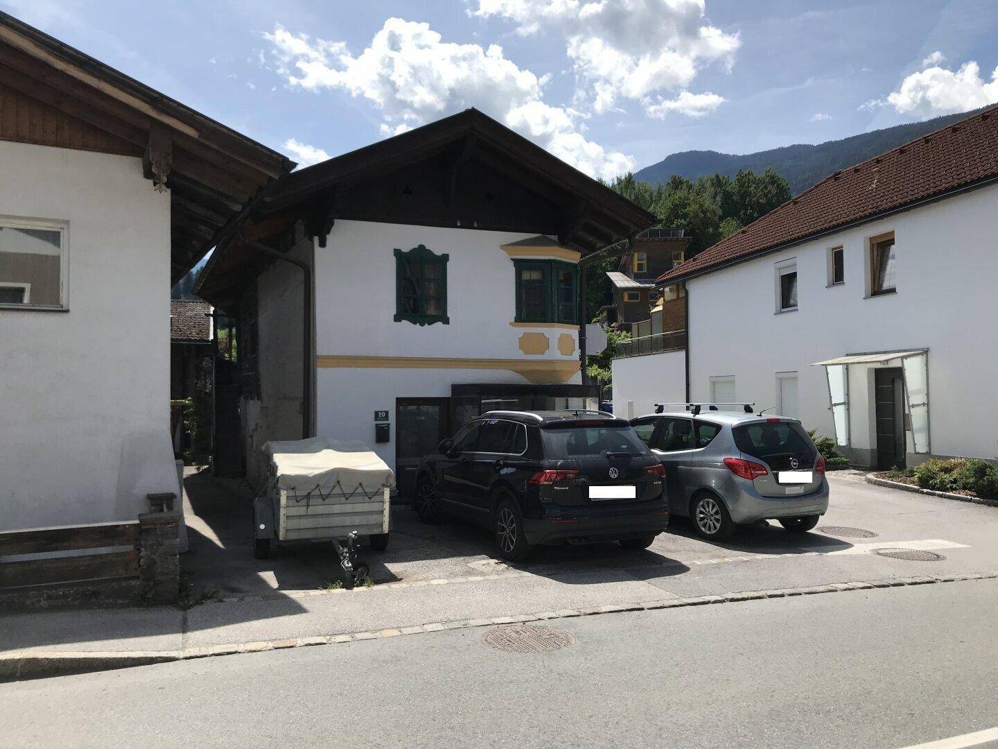 Traditionelles Wohnhaus mit geparkten Fahrzeugen und Bergblick in einer ländlichen Umgebung.