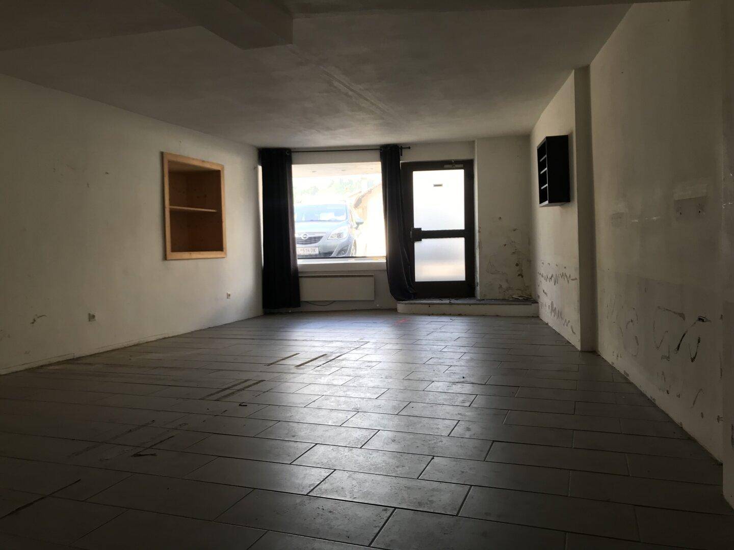 Großer, leerer Raum mit dunklen Fliesen und einem Fenster zur Straße.