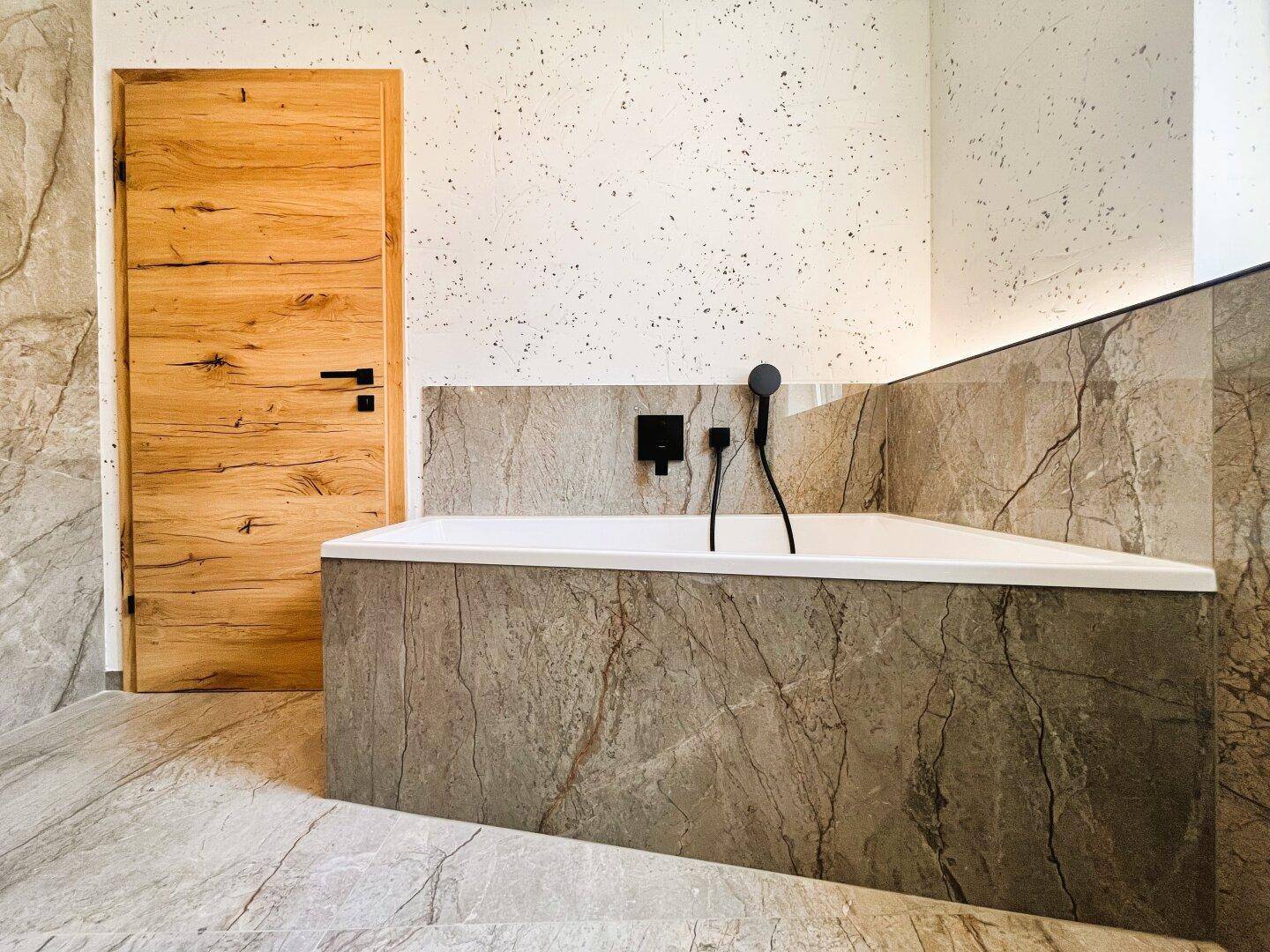Modernes Badezimmer mit eingebauter Badewanne und eleganten Marmorfliesen, Holztür sichtbar.