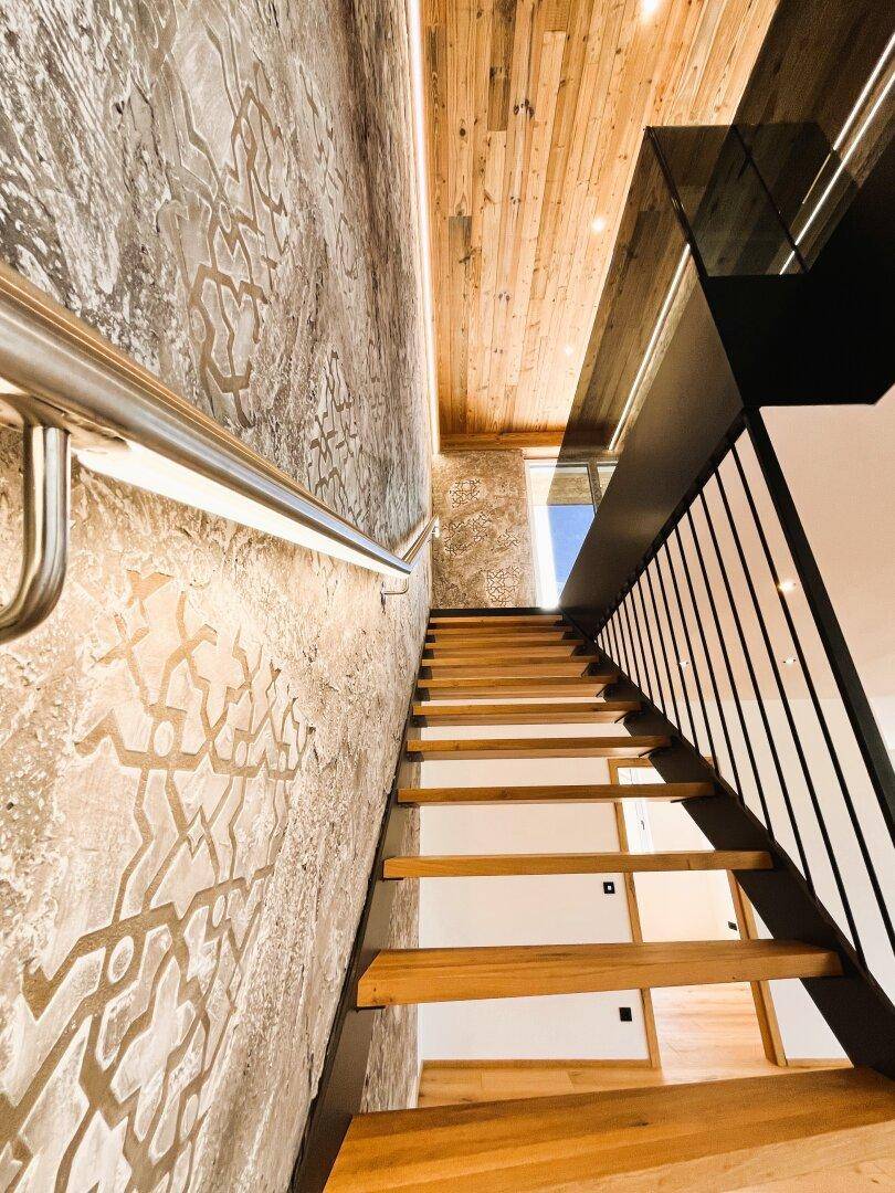 Aufgang einer modernen Holztreppe mit dekorativer Wand und Holzdecke, beleuchtet durch LED-Streifen.