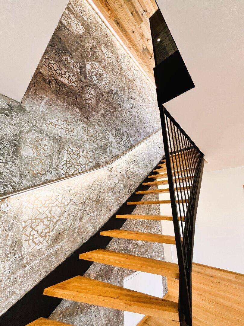 Elegante Holztreppe mit schwarzem Geländer und beleuchteter, gemusterter Wandverkleidung.
