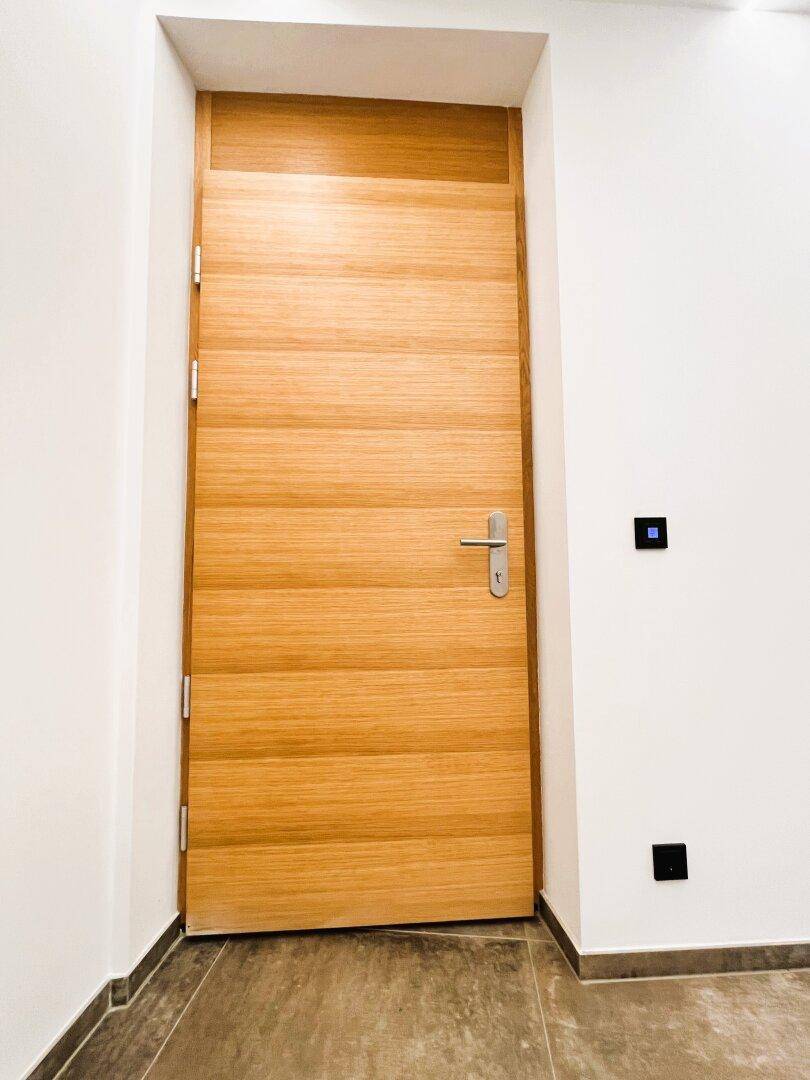 Moderne Holztür in einem hellen Flur mit dunklem Fliesenboden und weißen Wänden.