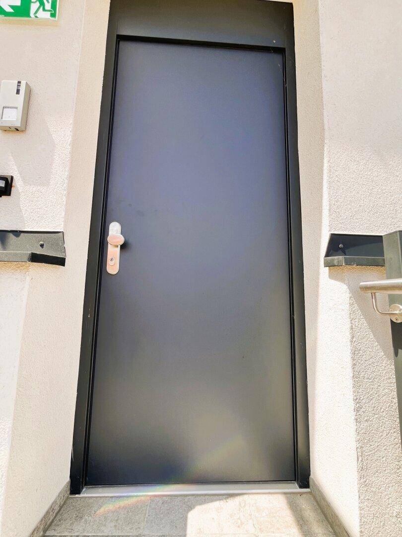Schwarze Eingangstür an einer hellen Fassade mit einem Bewegungsmelder darüber.