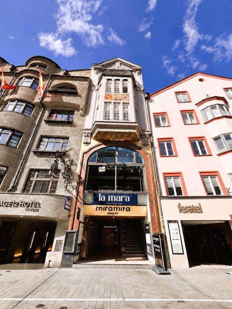 Historische Gebäudefassade in einer belebten Straße mit Geschäften im Erdgeschoss unter blauem Himmel.