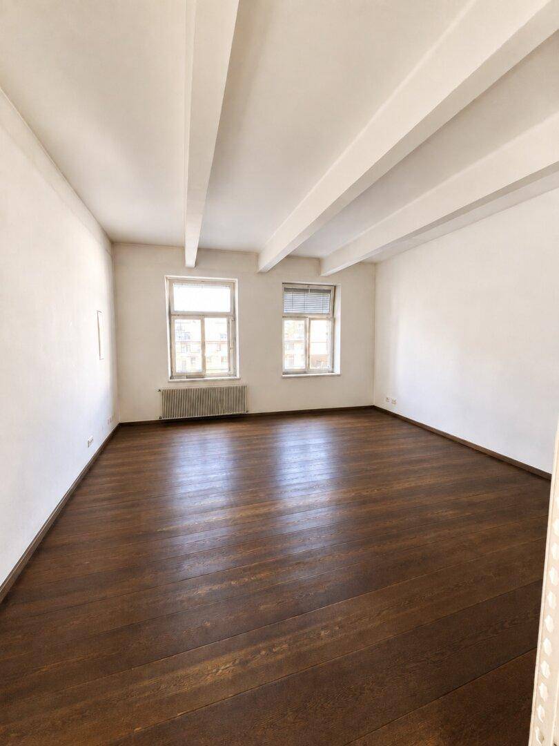Leerer Raum mit dunklem Holzboden, weißen Wänden und zwei Fenstern, die viel Tageslicht hereinlassen.