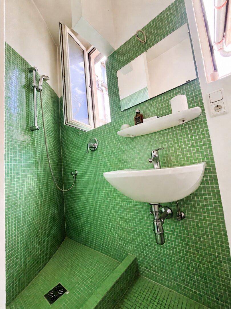 Kleines Badezimmer mit grünen Mosaikfliesen, Dusche, Waschbecken und einem Fenster für natürliche Belüftung.