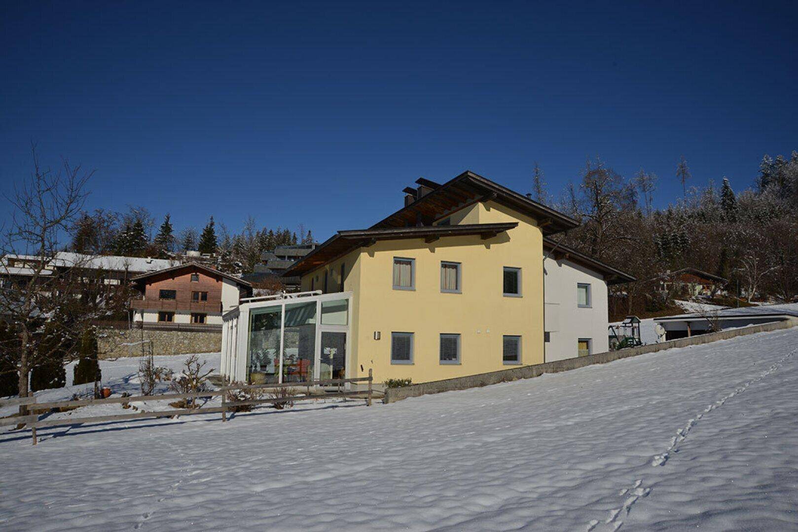 Hausansicht im Winter mit schneebedeckter Landschaft, gelber Fassade und großem Wintergarten.