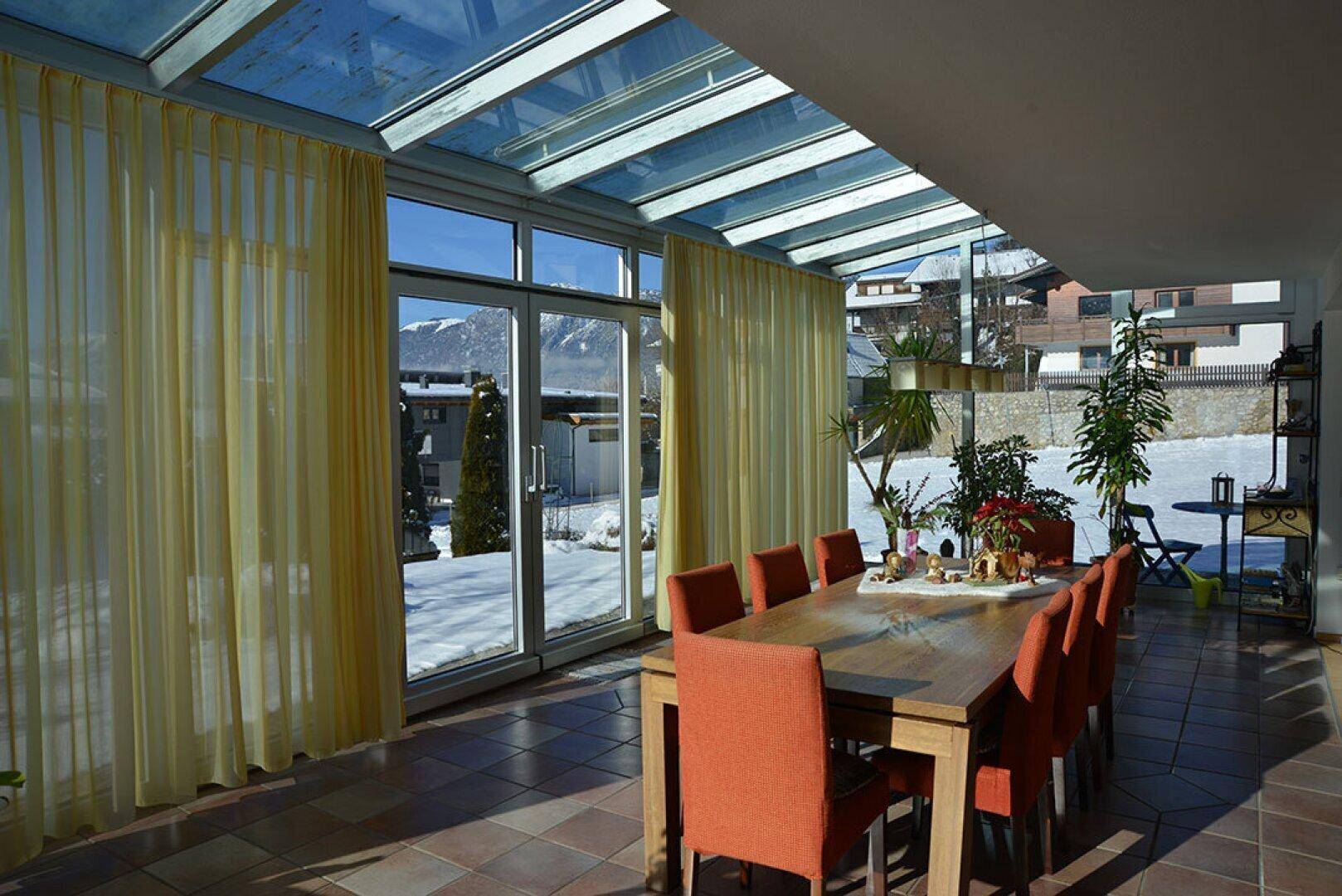 Heller Wintergarten mit großem Esstisch, Fliesenboden und Blick auf die schneebedeckte Landschaft.