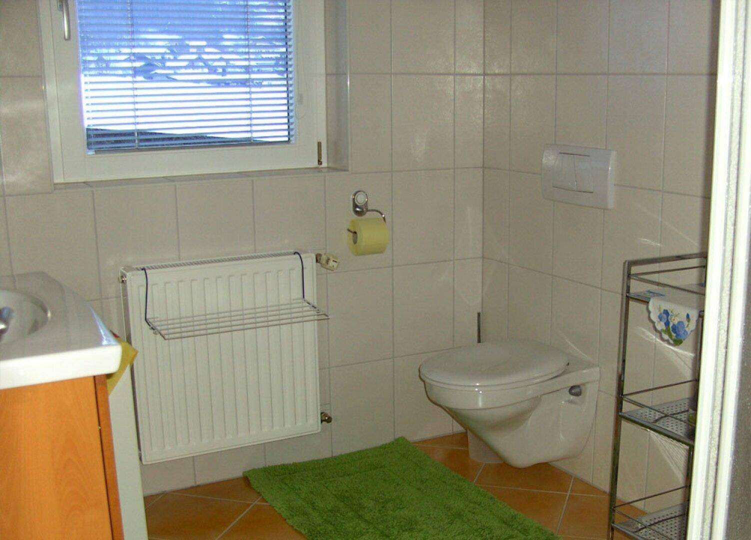 Helles Badezimmer mit weißen Fliesen, Toilette, Waschbecken und Fenster mit Jalousien.