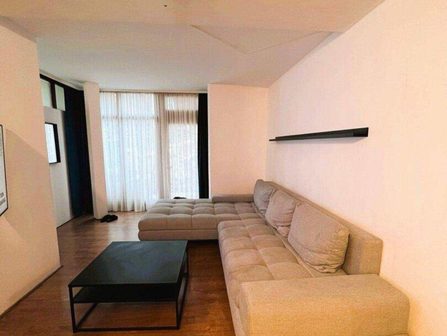 Geräumiges Wohnzimmer mit beigem Ecksofa, dunklem Couchtisch und viel Tageslicht.