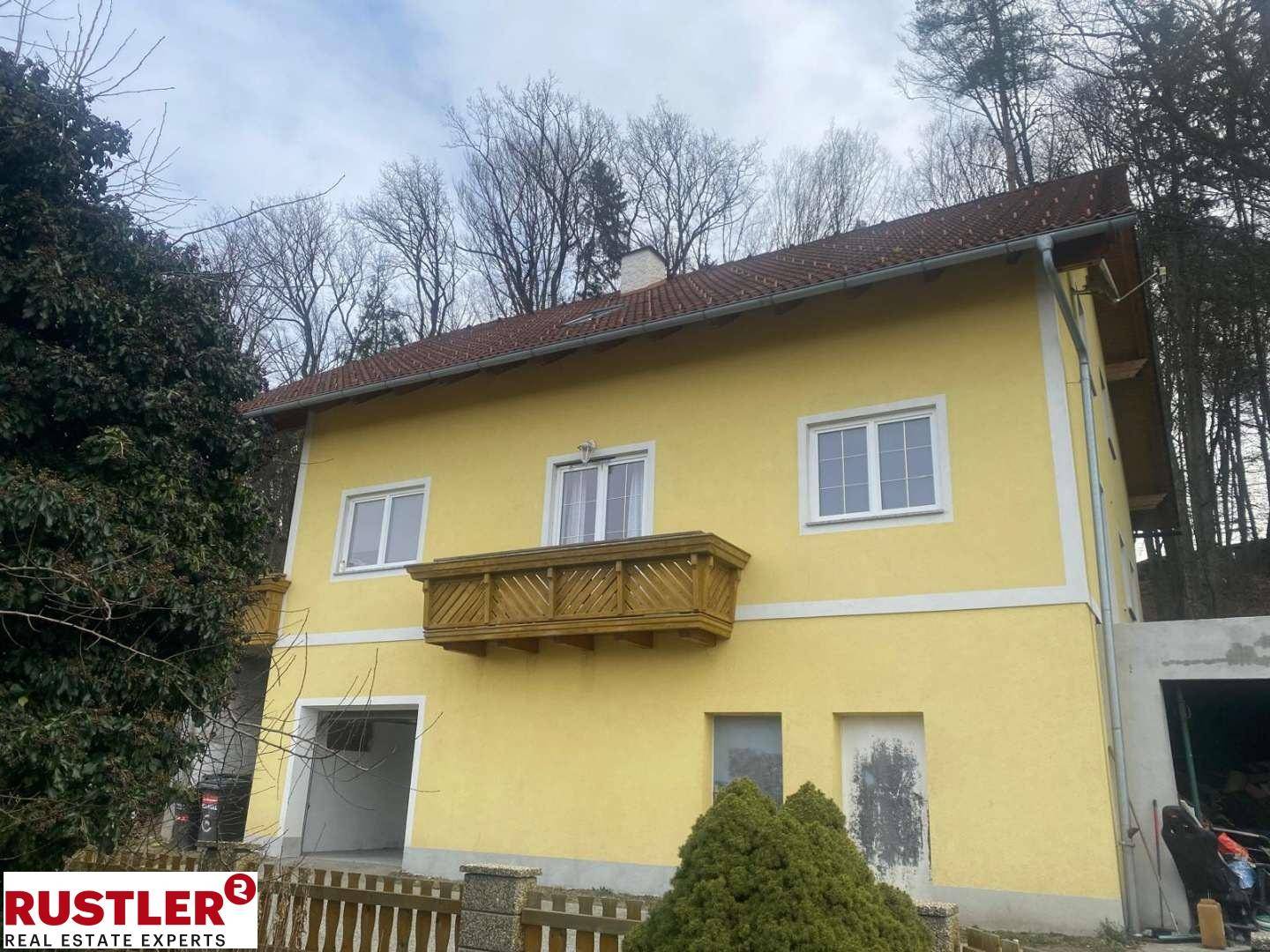 Ansicht des gelben Hauses mit Holzbalkon und Gartenbereich im Vordergrund.