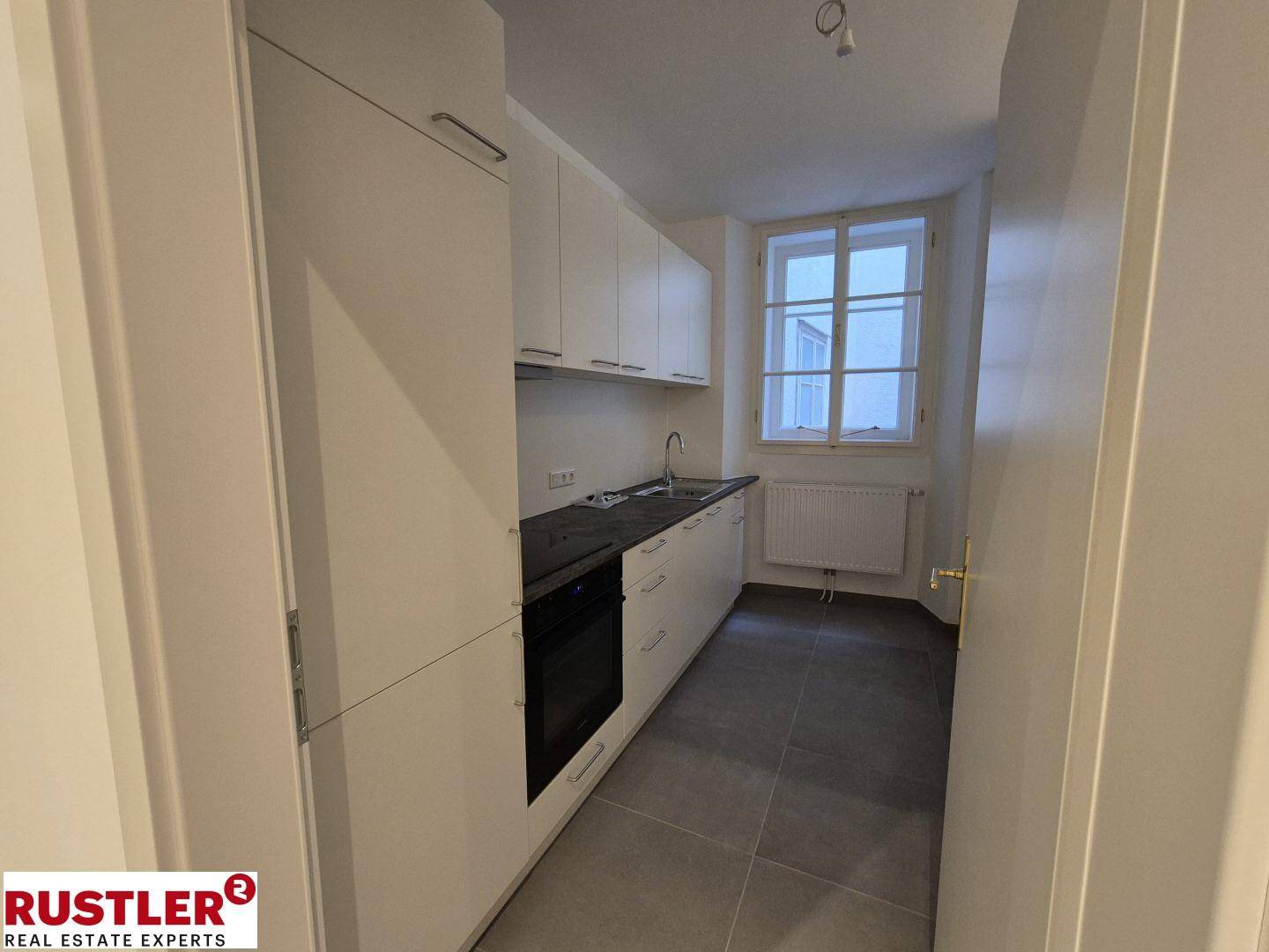 Moderne, schmale Küche mit weißen Einbauschränken, dunkler Arbeitsplatte und Fenster.