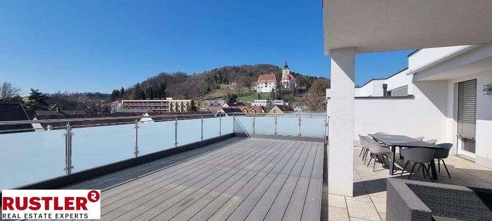 Weitläufige Terrasse mit Glasgeländer und Panoramablick auf die umliegende Landschaft und einen Hügel mit Kirche.