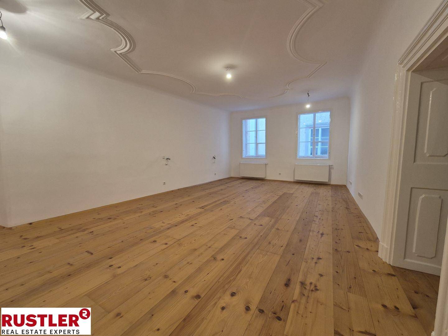 Geräumiges Wohnzimmer mit hellem Holzfußboden, hohen Decken und Stuckverzierungen an der Decke.