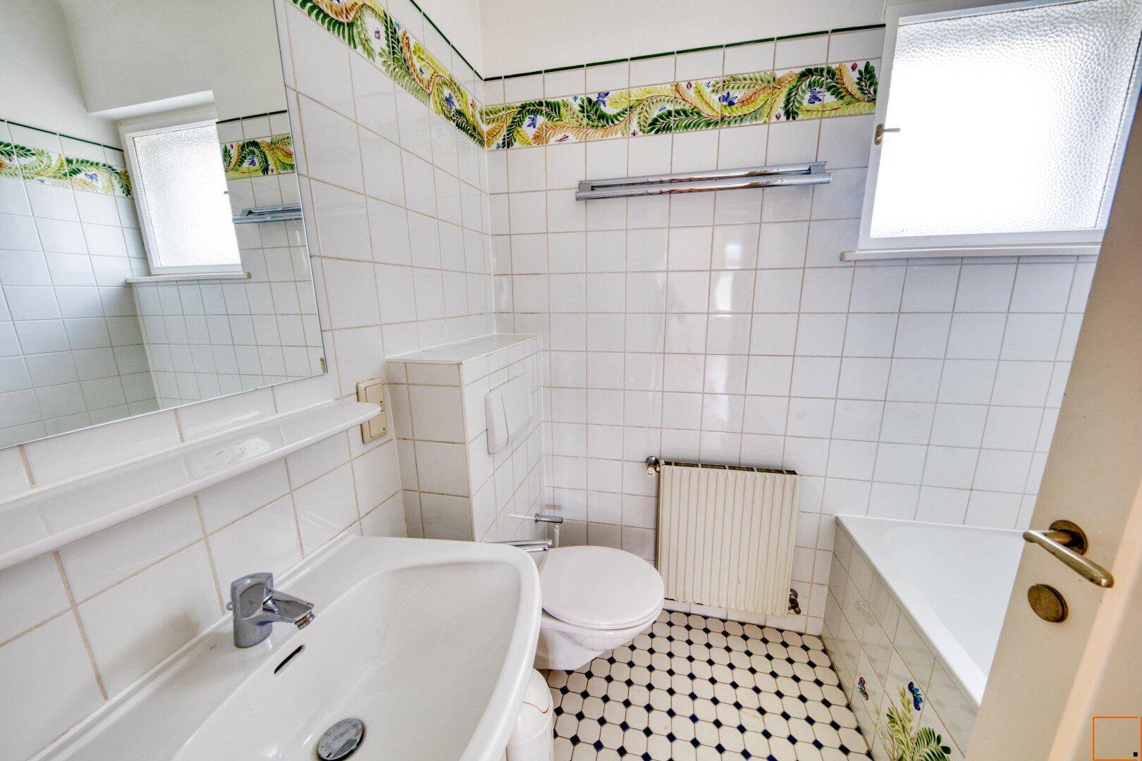 Badezimmer mit weißen Fliesen, Fenster und einem dekorativen floralen Fliesenband.