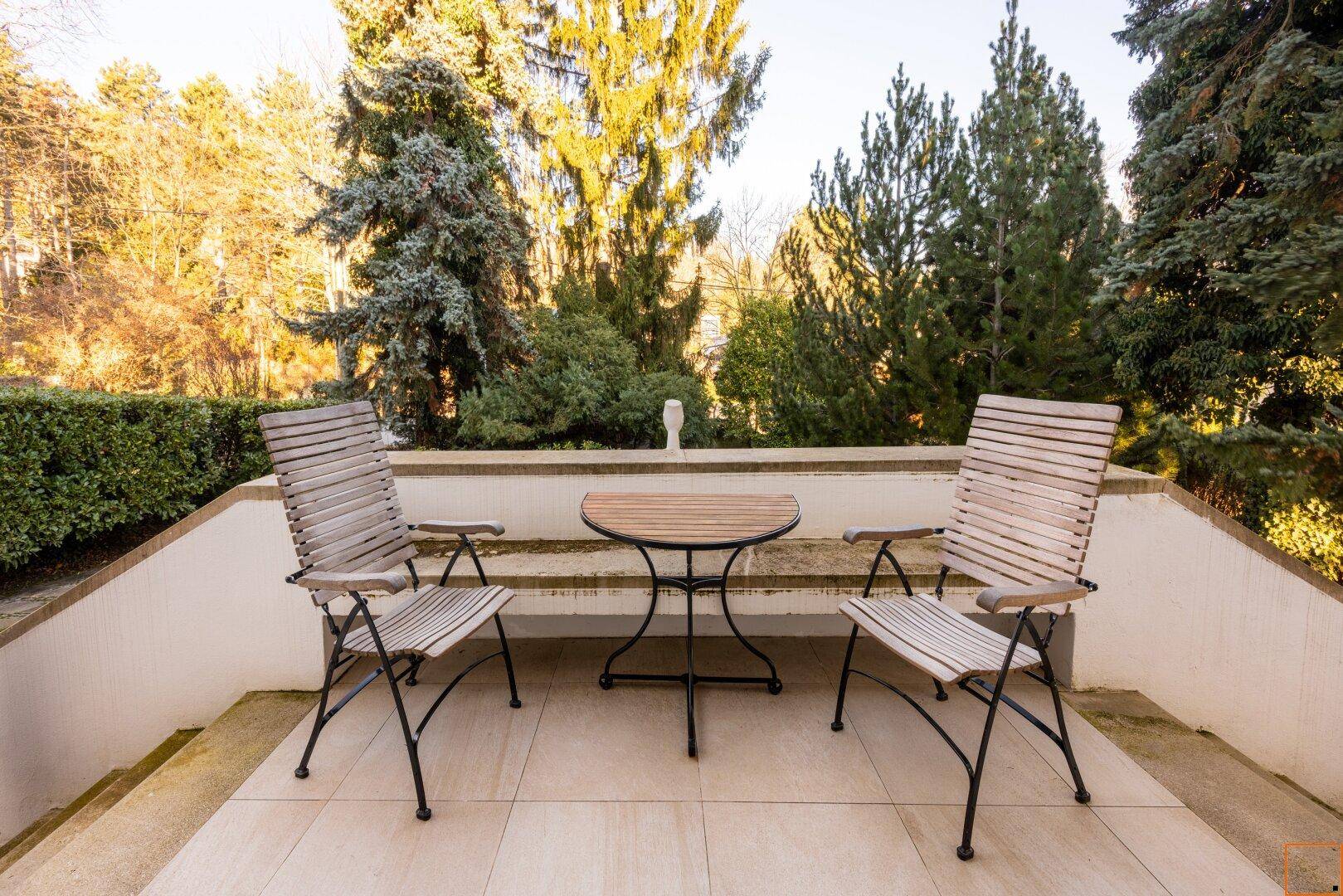 Gemütlicher Sitzbereich im Freien auf einer Terrasse mit Tisch und Stühlen, umgeben von Grün.